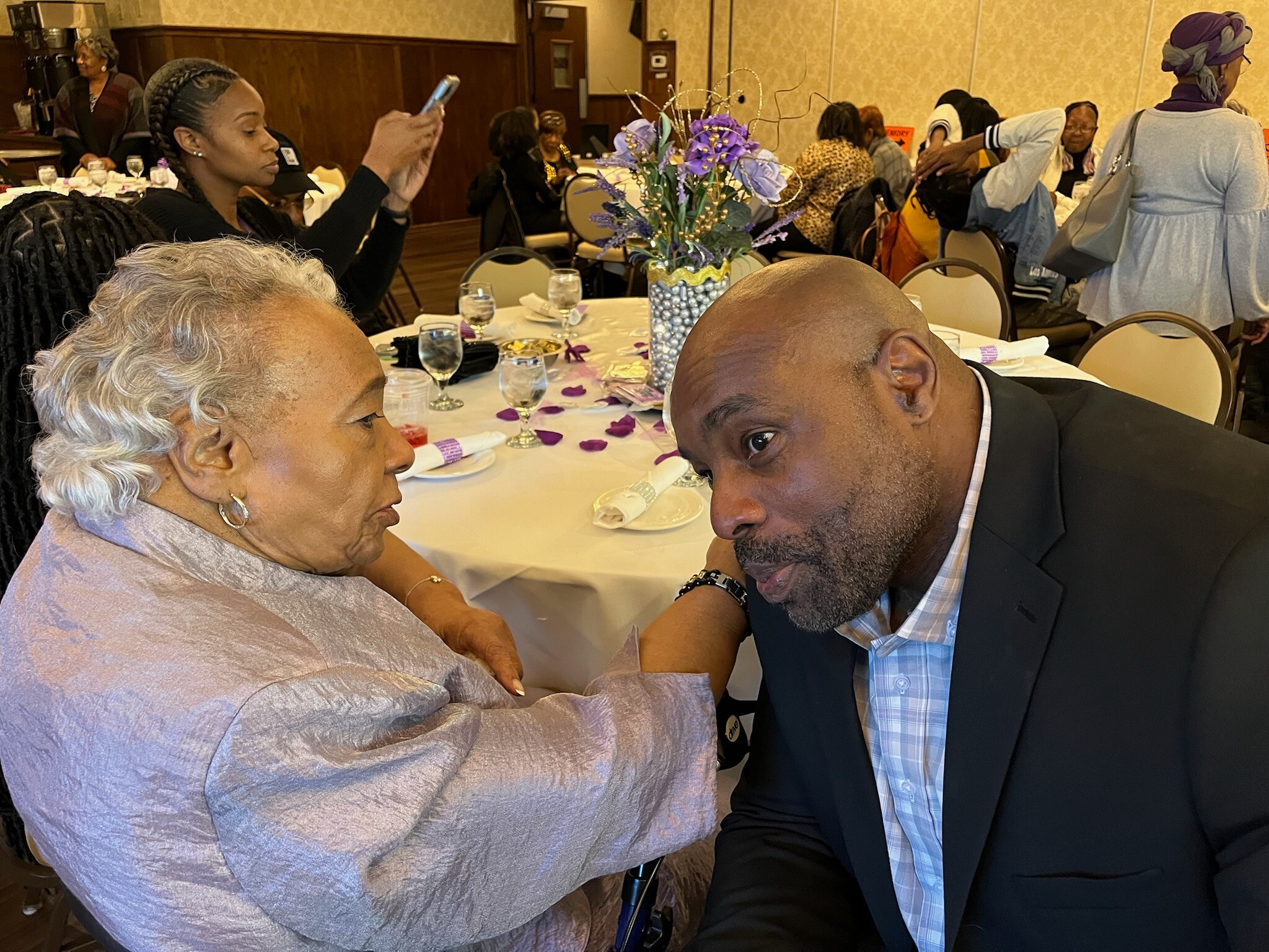 Two elderly adults lean in toward each other with foreheads touching at a banquet table during a celebration.