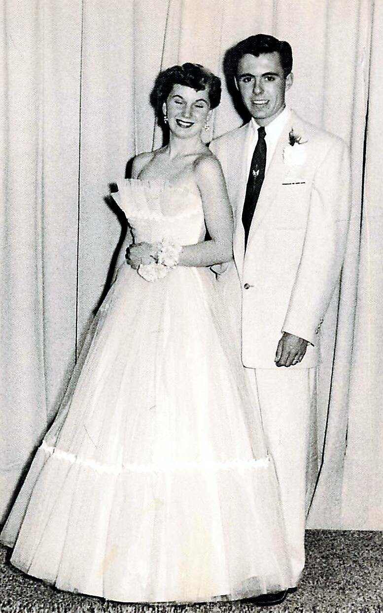 Bride and groom posing on their wedding day; she wears a white strapless gown and holds a bouquet, he stands beside in a light suit with a dark tie.