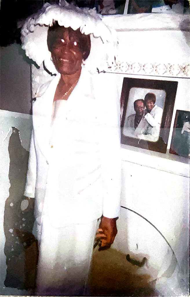 Smiling elderly woman in a white suit and large white hat, standing indoors beside a shelf with framed photos.