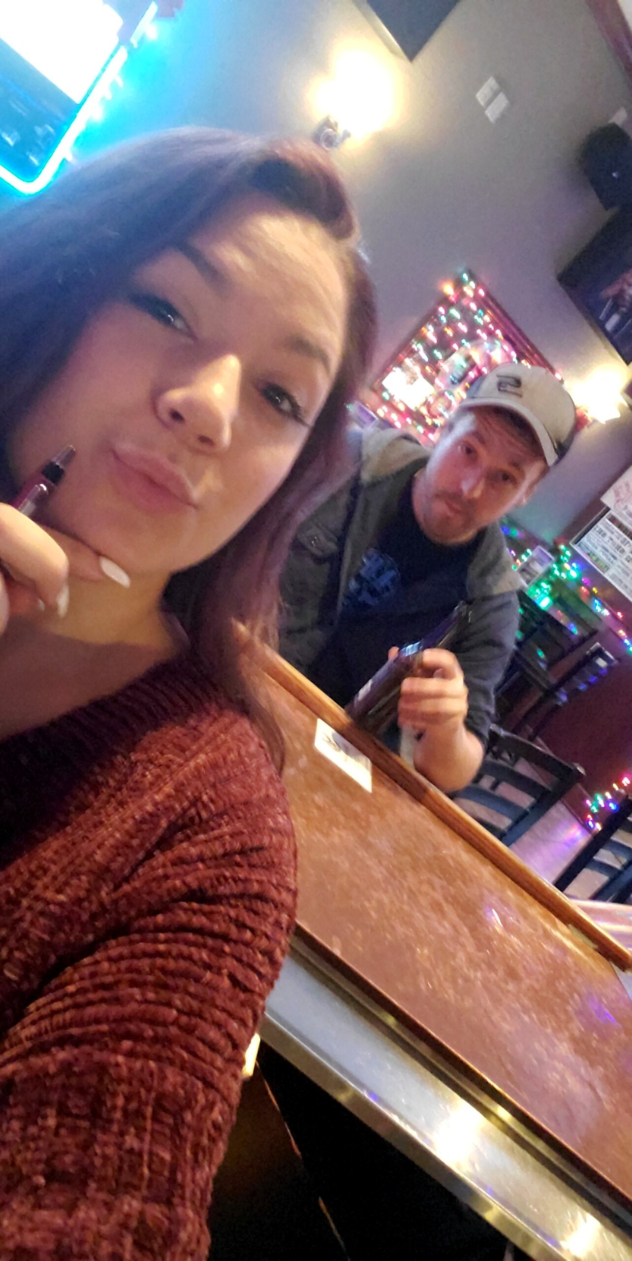 Woman in a maroon sweater takes a selfie at a neon-lit bar, with a man in a cap holding a bottle in the background.
