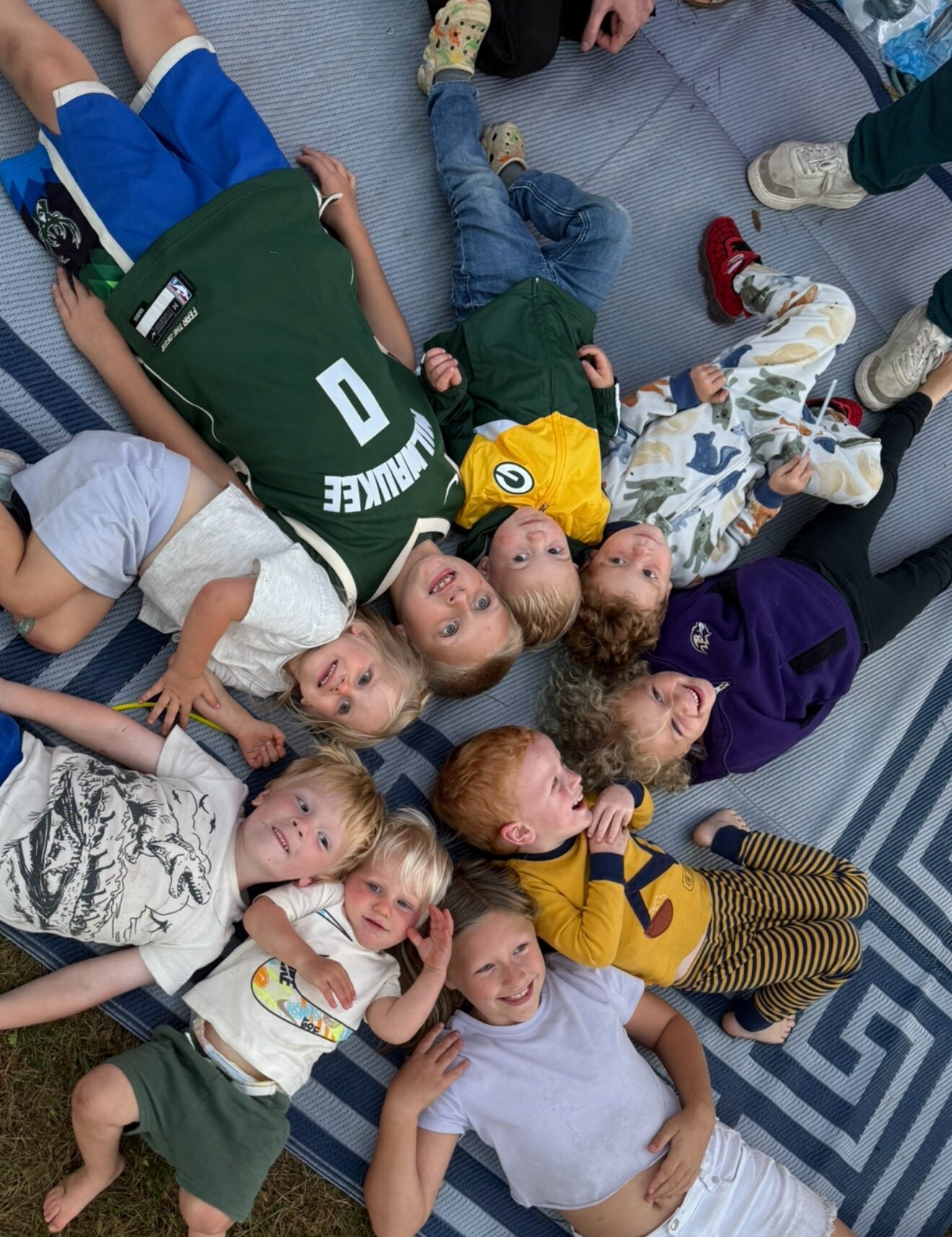 A group of smiling children lying on a blue striped blanket outdoors, arranged in a circle and looking up at the camera.