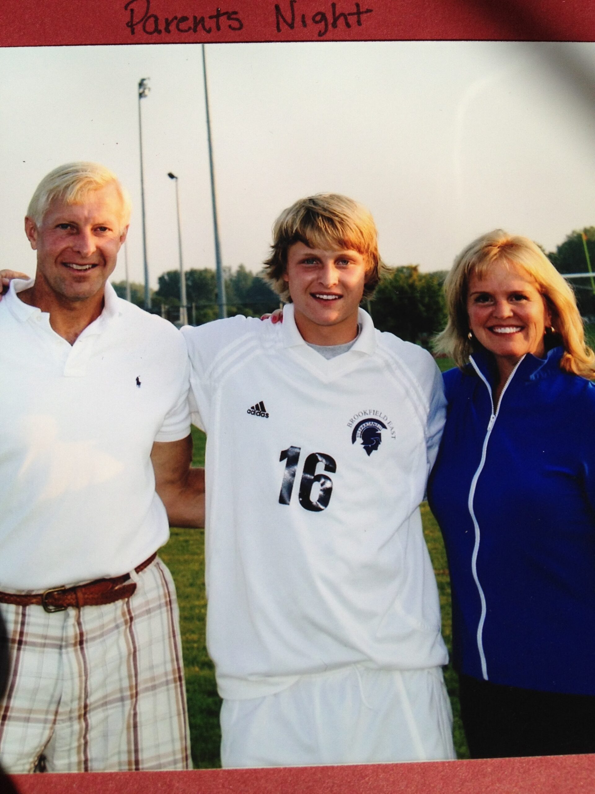 Three people pose together outdoors after a sports event: an man in a white polo, a young athlete in a white jersey with number 16, and a smiling woman in a blue jacket, with a 'Parents Night' banner above.
