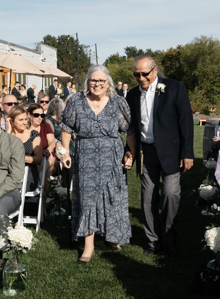 Smiling elderly couple walks hand in hand down an outdoor wedding aisle: woman in a blue patterned dress with a corsage, man in a dark blazer with a boutonniere; guests sit on either side on a sunny lawn.