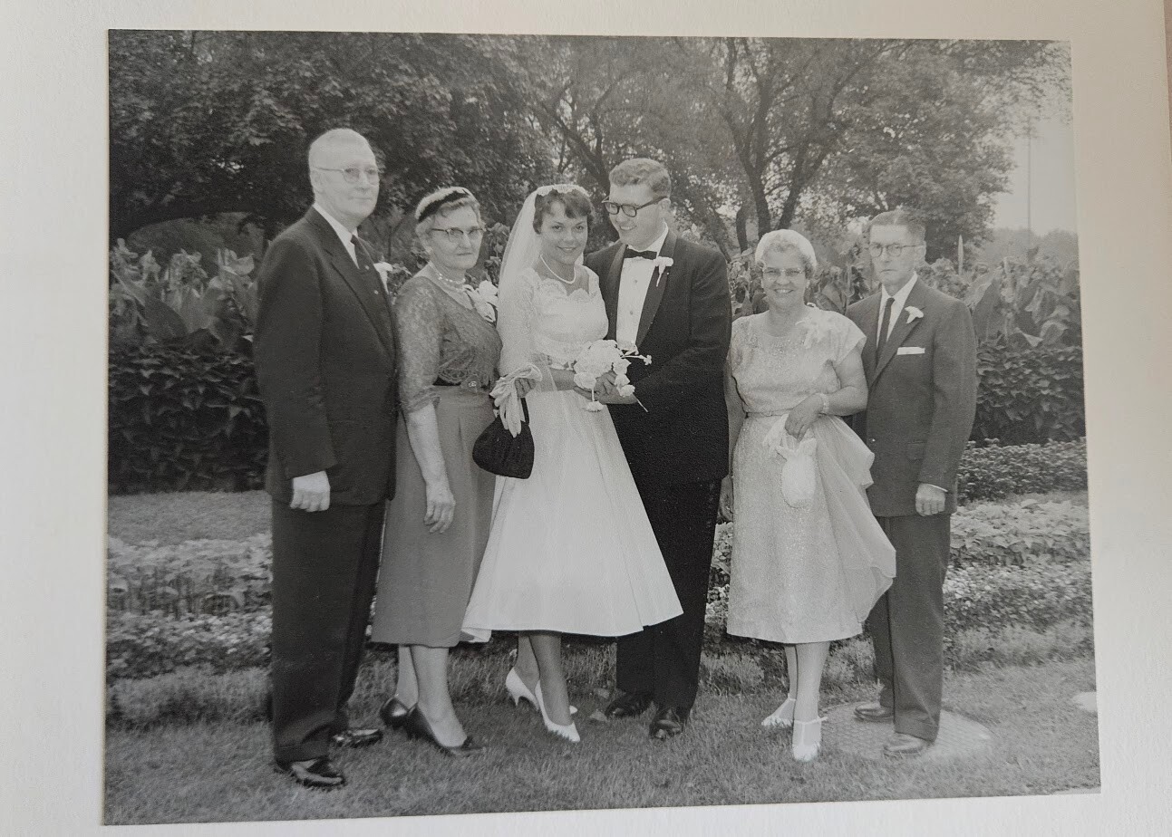 Bride and groom stand center in wedding attire, flanked by two couples outdoors in a garden setting.