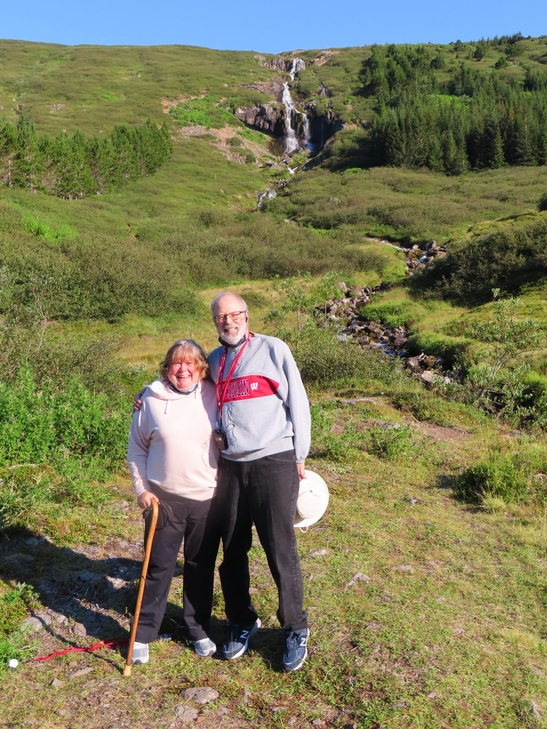 Smiling older couple posing on a rocky hillside with a green valley and a distant waterfall behind them.