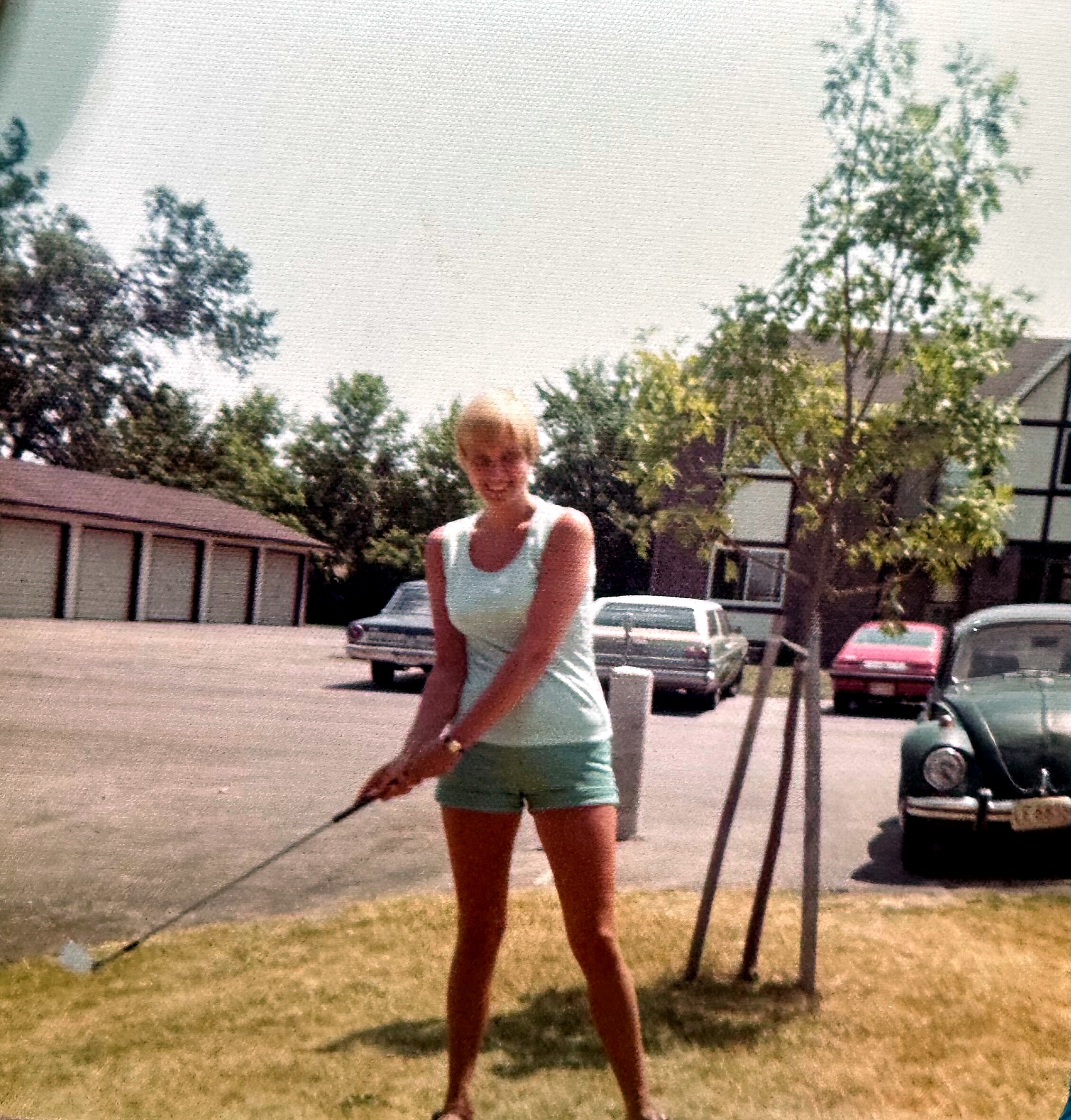 Woman in a light teal swimsuit stands in a suburban backyard with trees and parked cars in the background.
