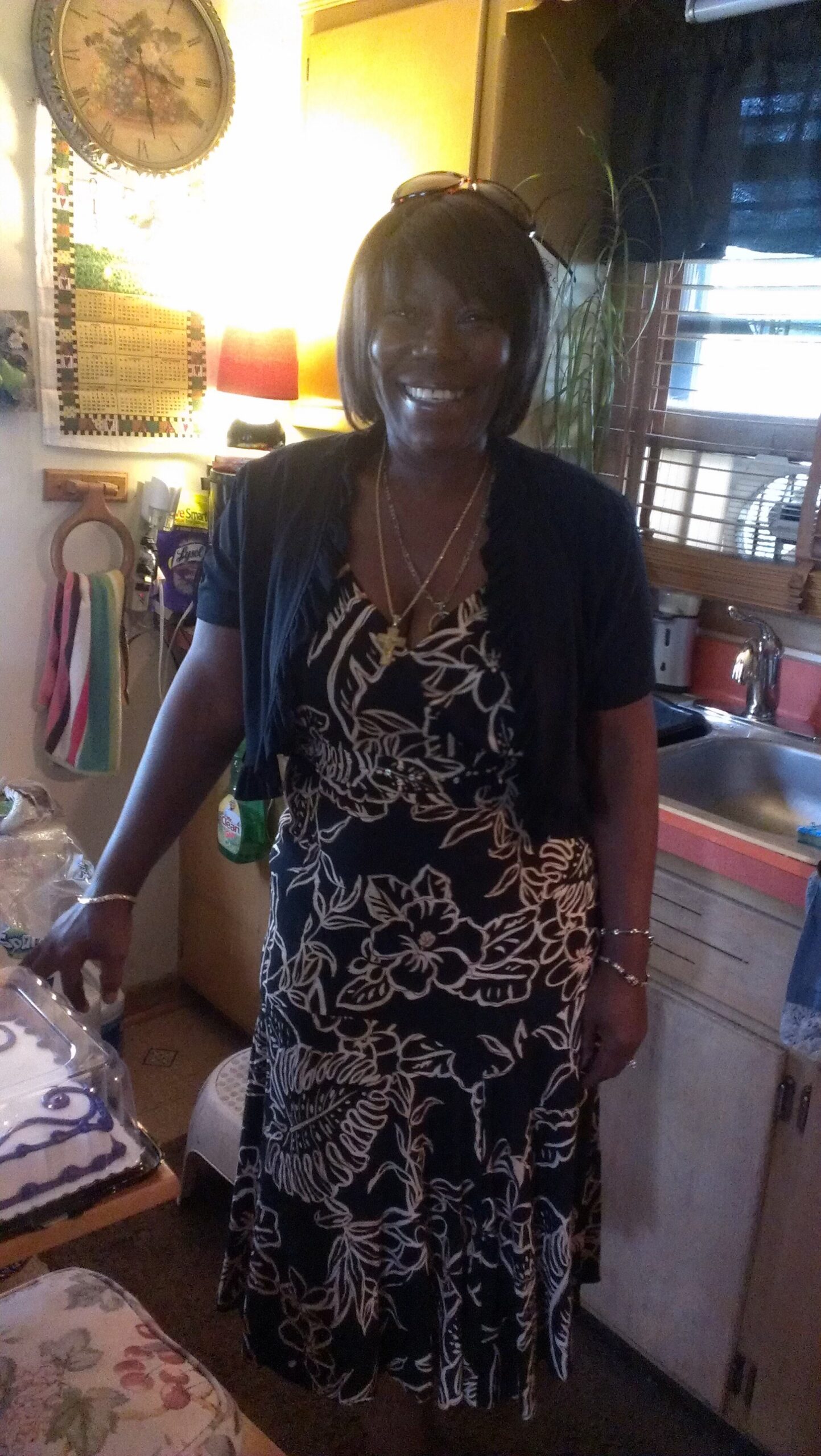 Smiling woman in a black-and-white floral maxi dress stands in a home kitchen, sunglasses on her head.