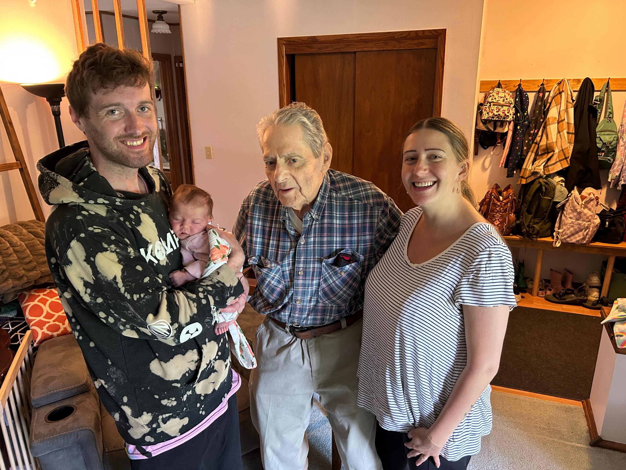 A man wearing a tie-dye hoodie cradles a sleeping baby as a man in plaid stands beside a smiling woman in a striped dress in a home hallway with coats on hooks in the background.