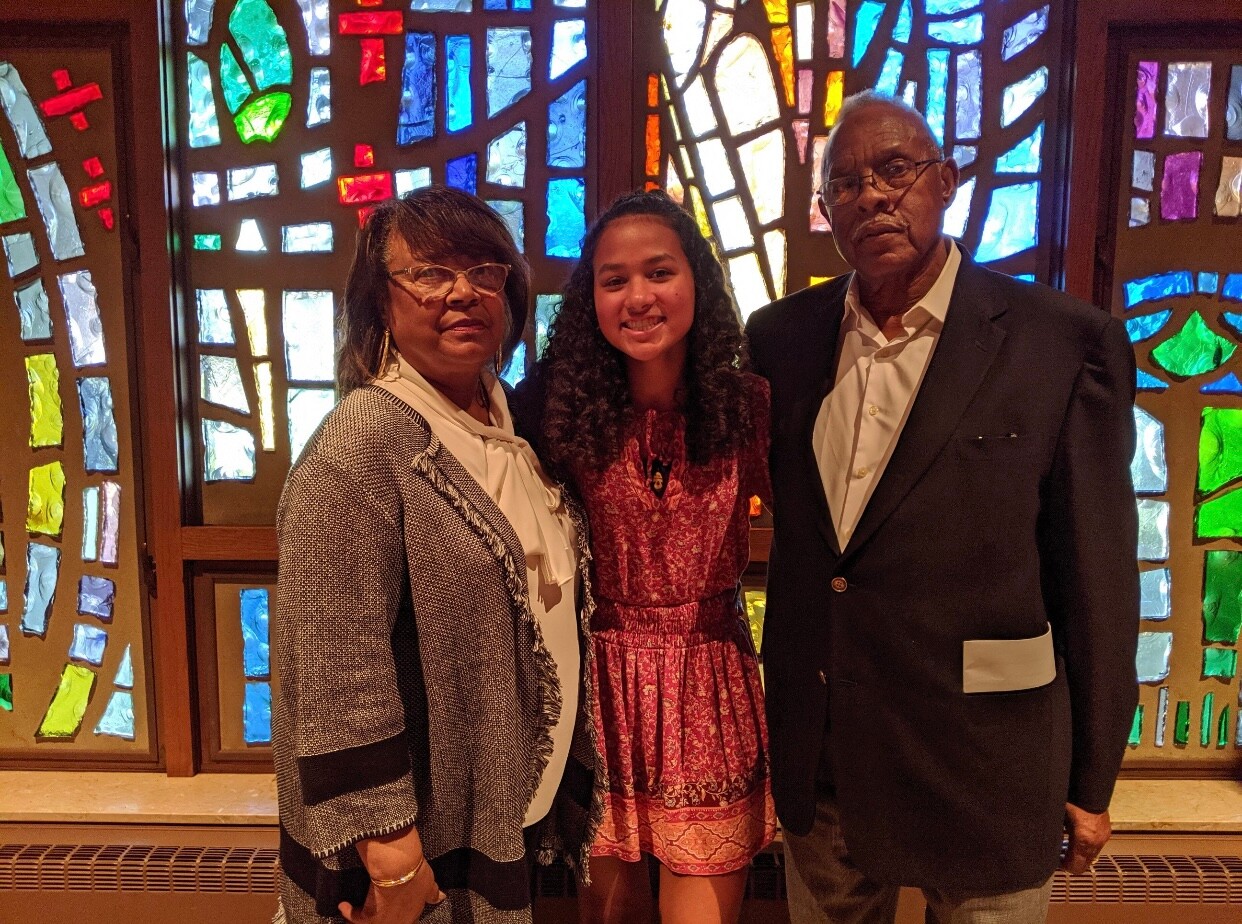 Three people posing together in front of a vibrant stained-glass window: a woman on the left, a girl in the middle, and a man on the right.