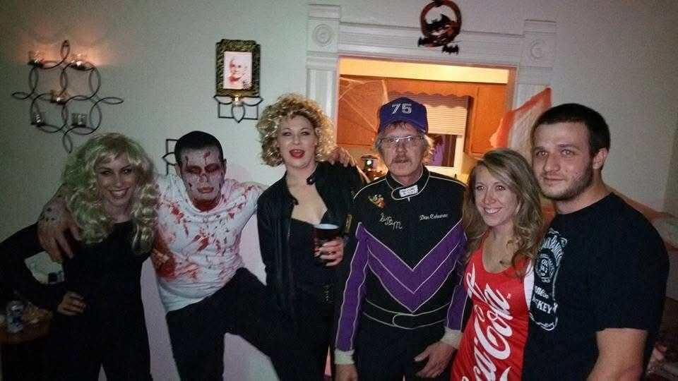 Six friends in a living room wearing Halloween costumes pose together for a photo, some with makeup and colorful outfits. - The group includes a zombie-like character, a blonde in black, and others smiling into the camera.