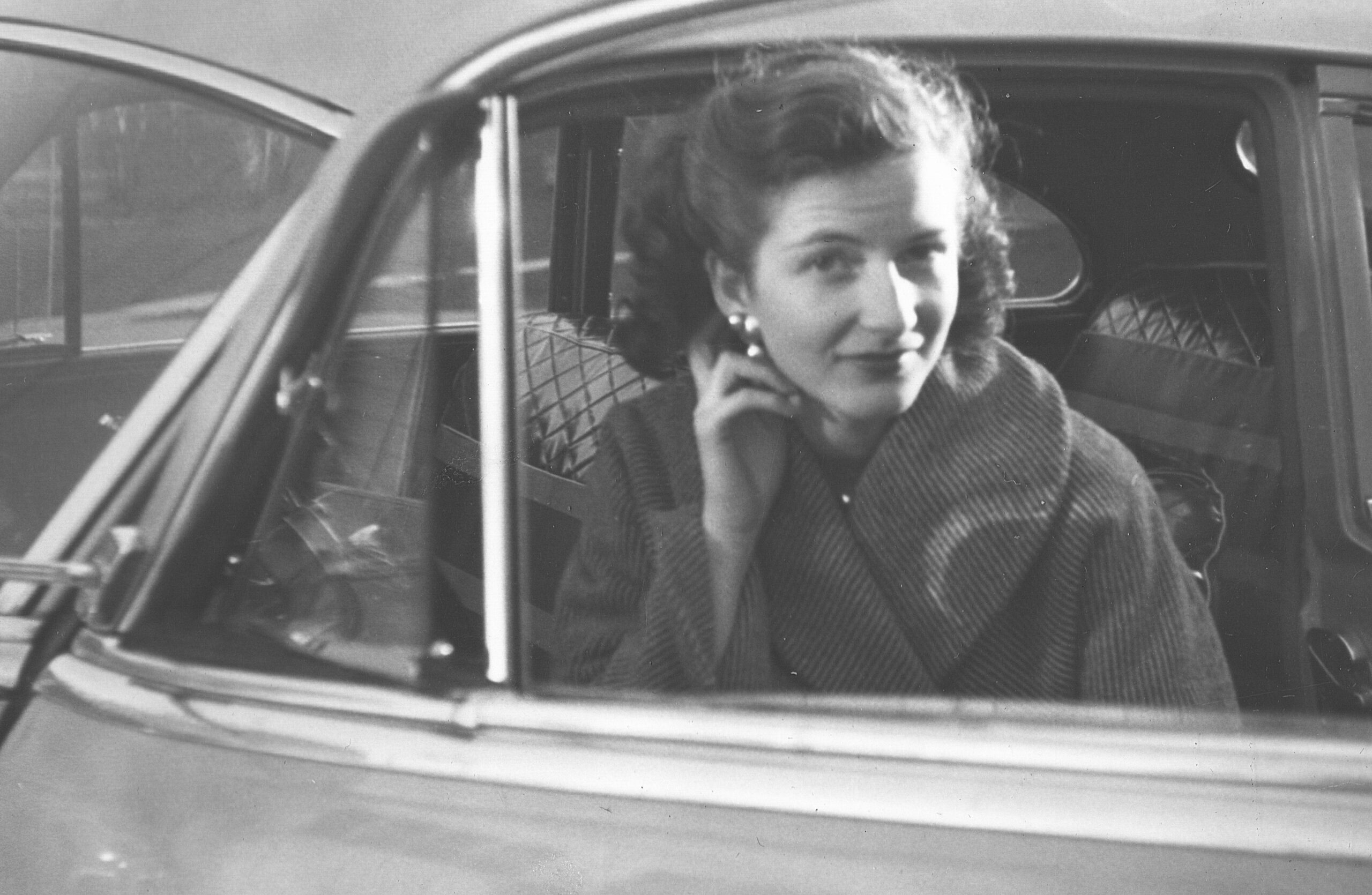 Woman in a plaid coat sits in a car and talks on a handheld phone, looking toward the camera (black and white photo).