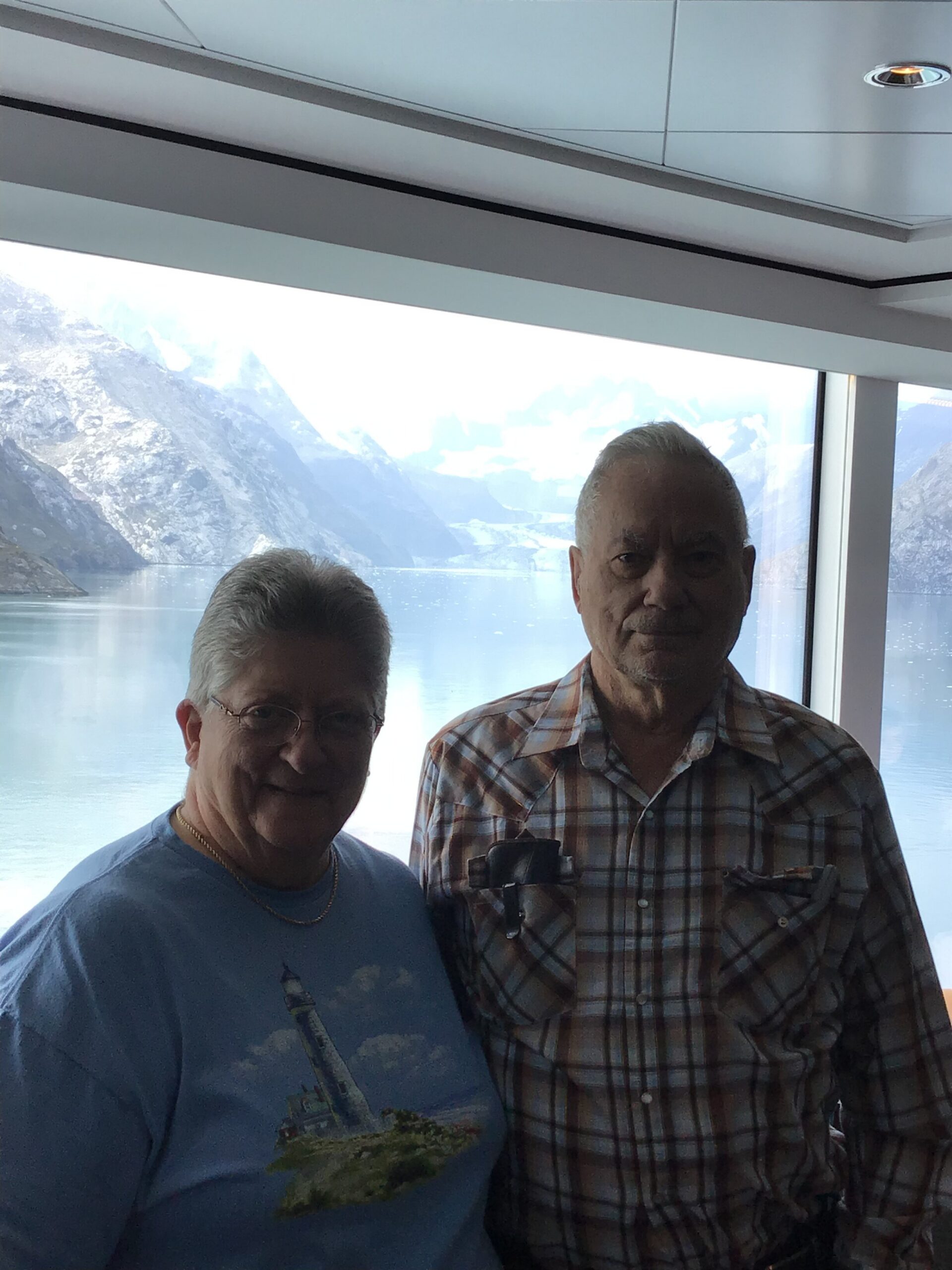 Two older men posing for a photo indoors, with snow-capped mountains and a lake visible through the window behind them.