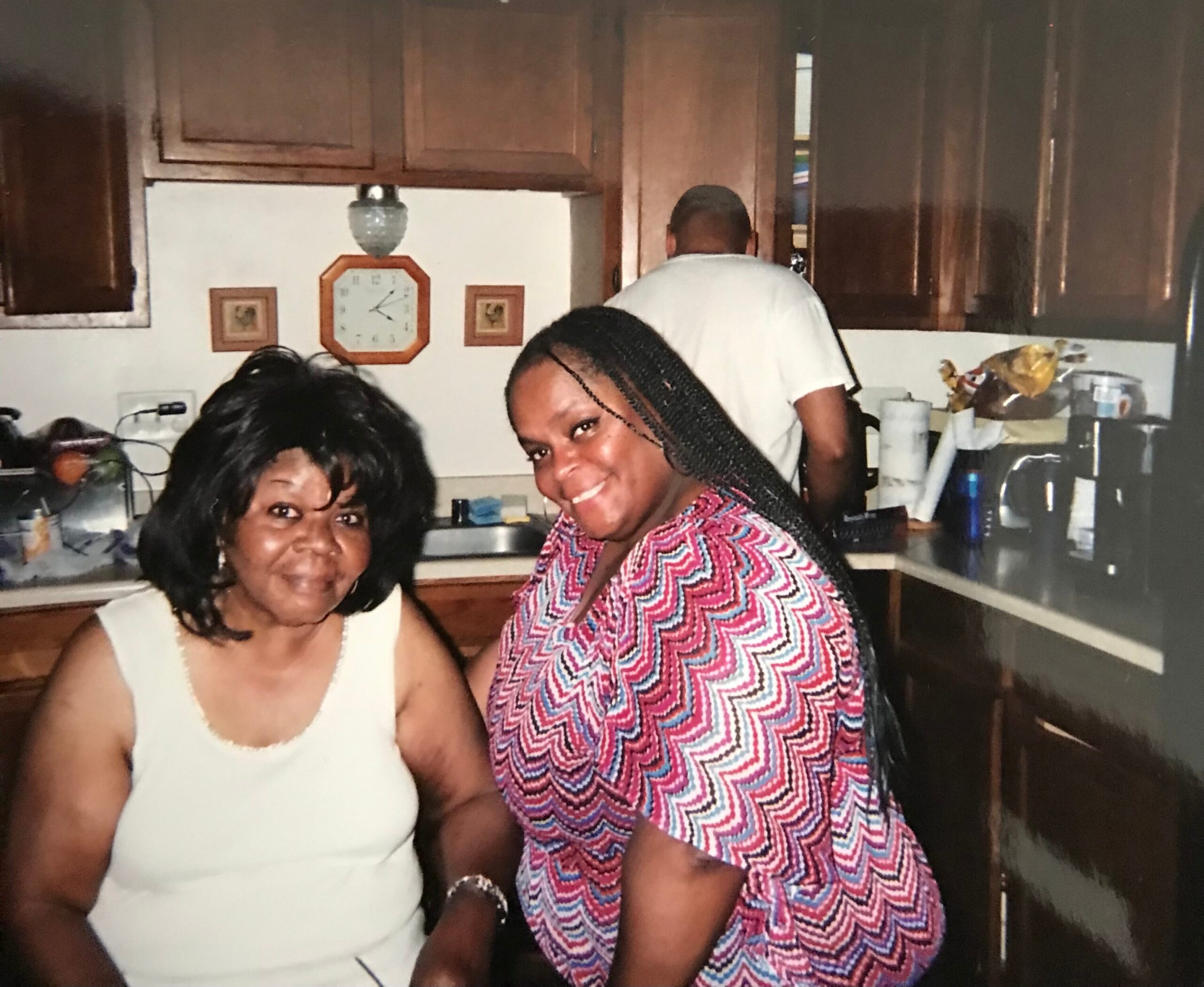 Two women smiling in a home kitchen with wooden cabinets and a wall clock behind them.
