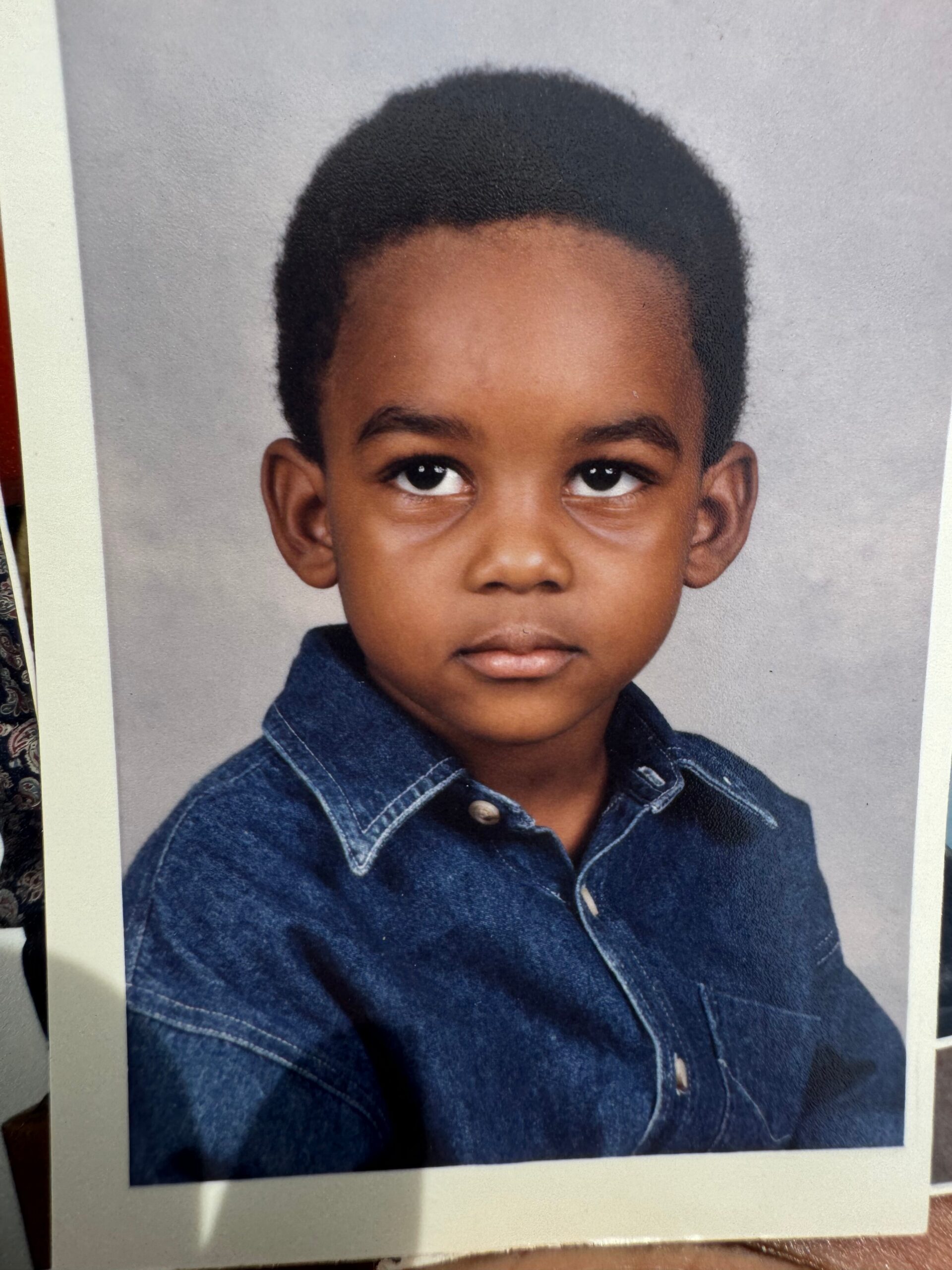 Close-up portrait of a young boy with short black hair wearing a blue denim shirt against a neutral gray background, looking forward with a calm expression.
