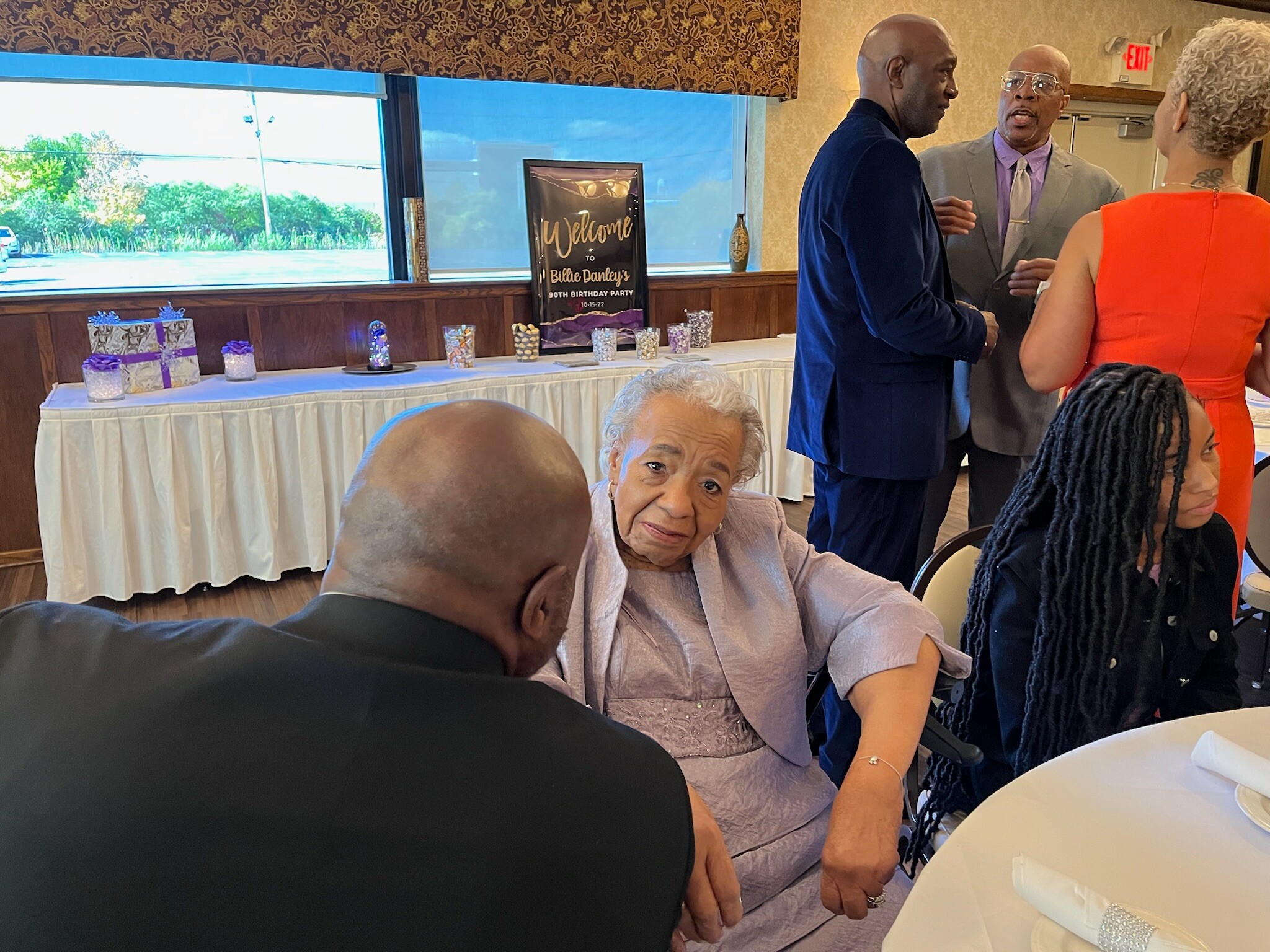 Senior woman in a lavender dress seated at a table during a birthday party, smiling as guests chat nearby