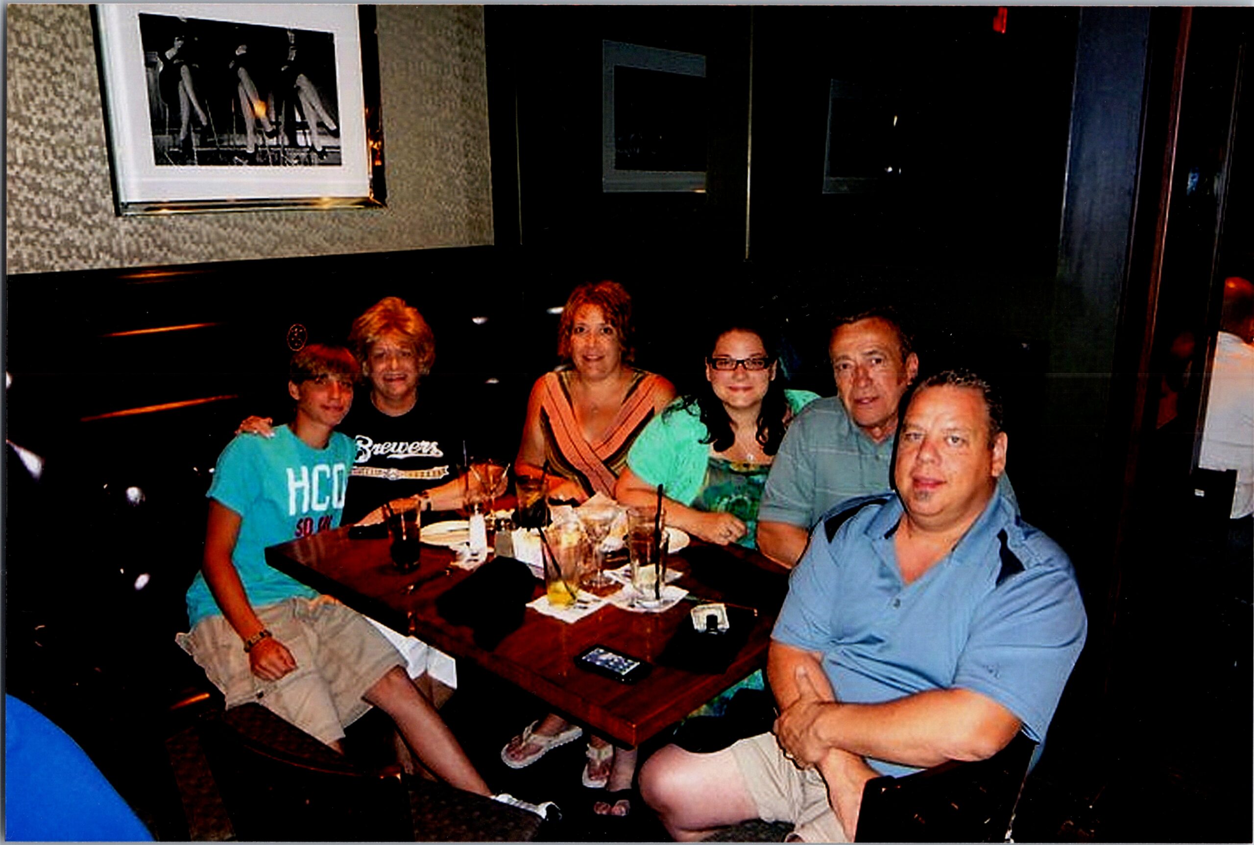Six people gathered around a restaurant table, smiling at the camera with drinks and plates in front of them in a dim setting.