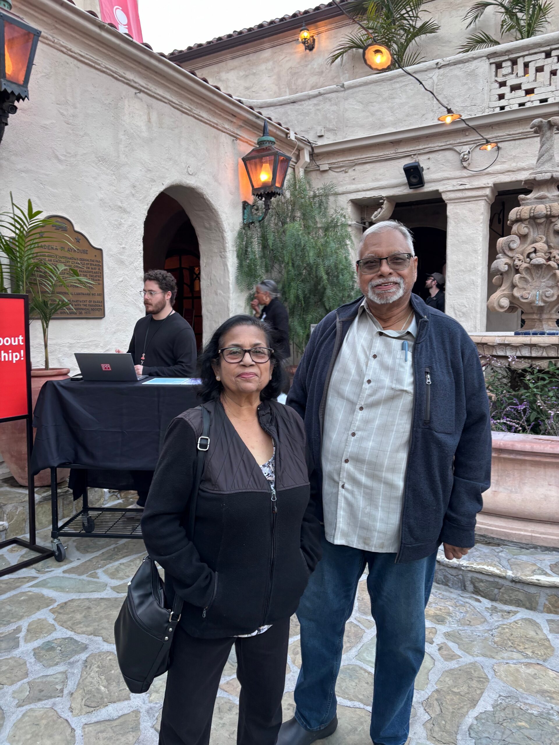 Smiling older couple posing in a sunny courtyard with a stone tile floor, a fountain sculpture, and string lights in the background.