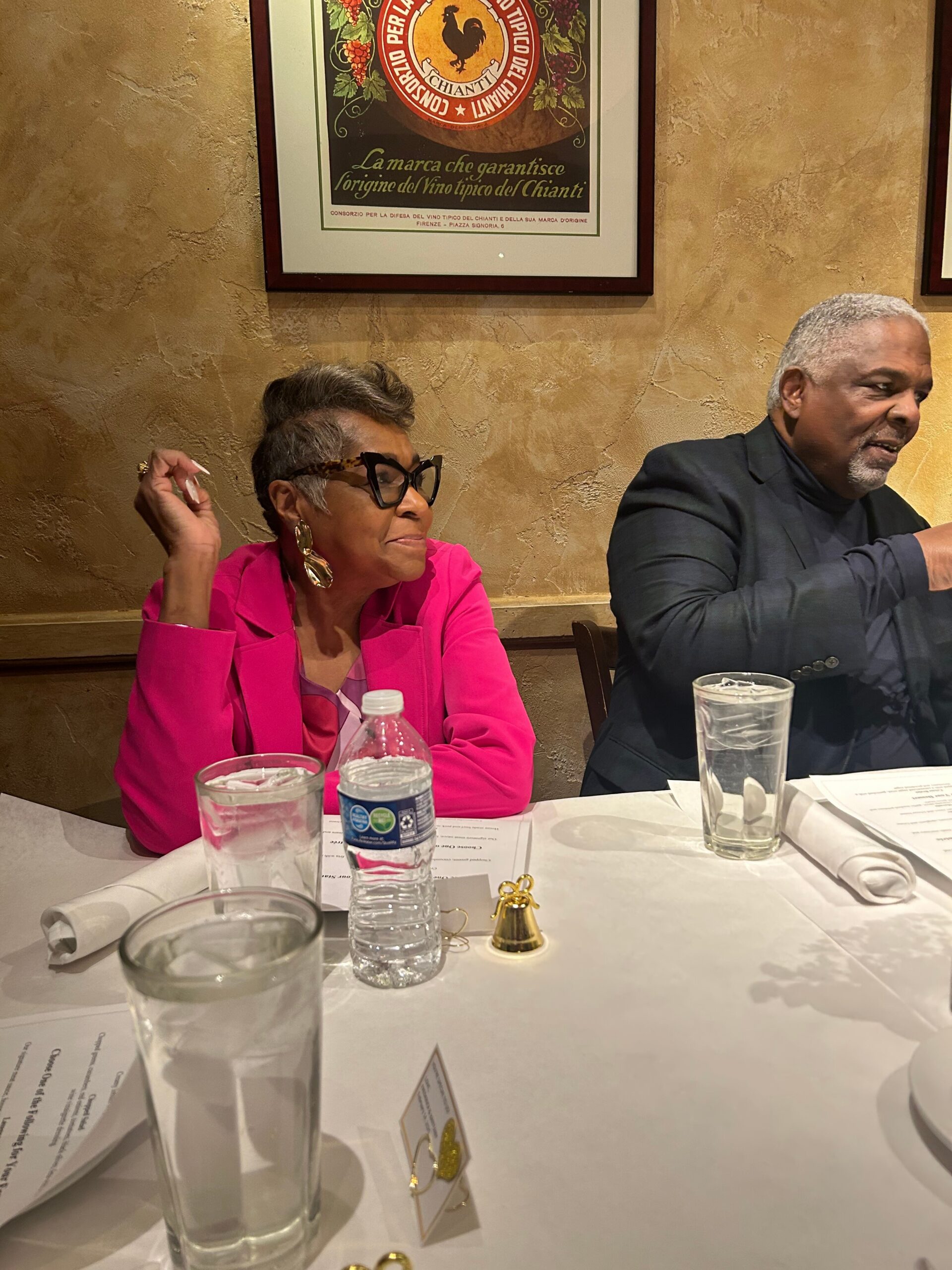 Woman in a bright pink blazer and large earrings sits at a restaurant table beside a man in a dark suit, with drinks and papers on the table.