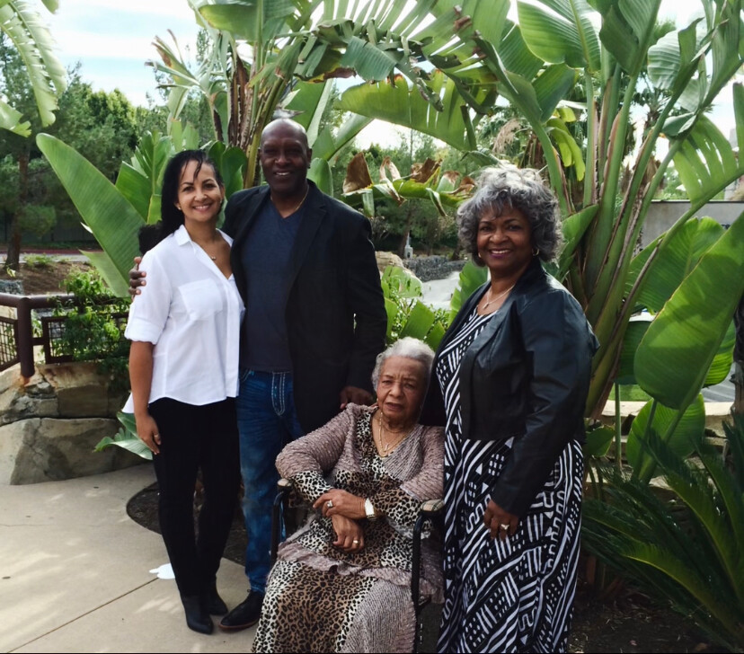 Four adults posing outdoors among large tropical plants; an elderly woman in a wheelchair sits center, flanked by a man and two women, all smiling.