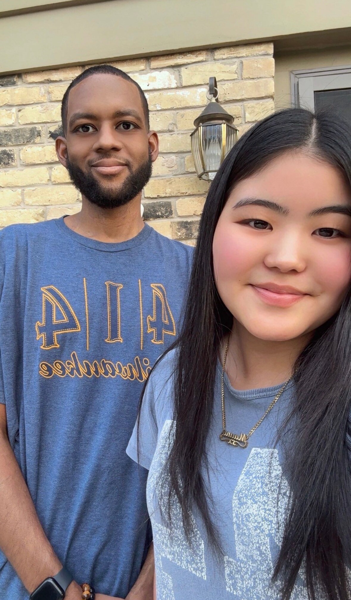 Close-up selfie of a man and a woman smiling outdoors in front of a brick wall with a hanging lantern light behind them.