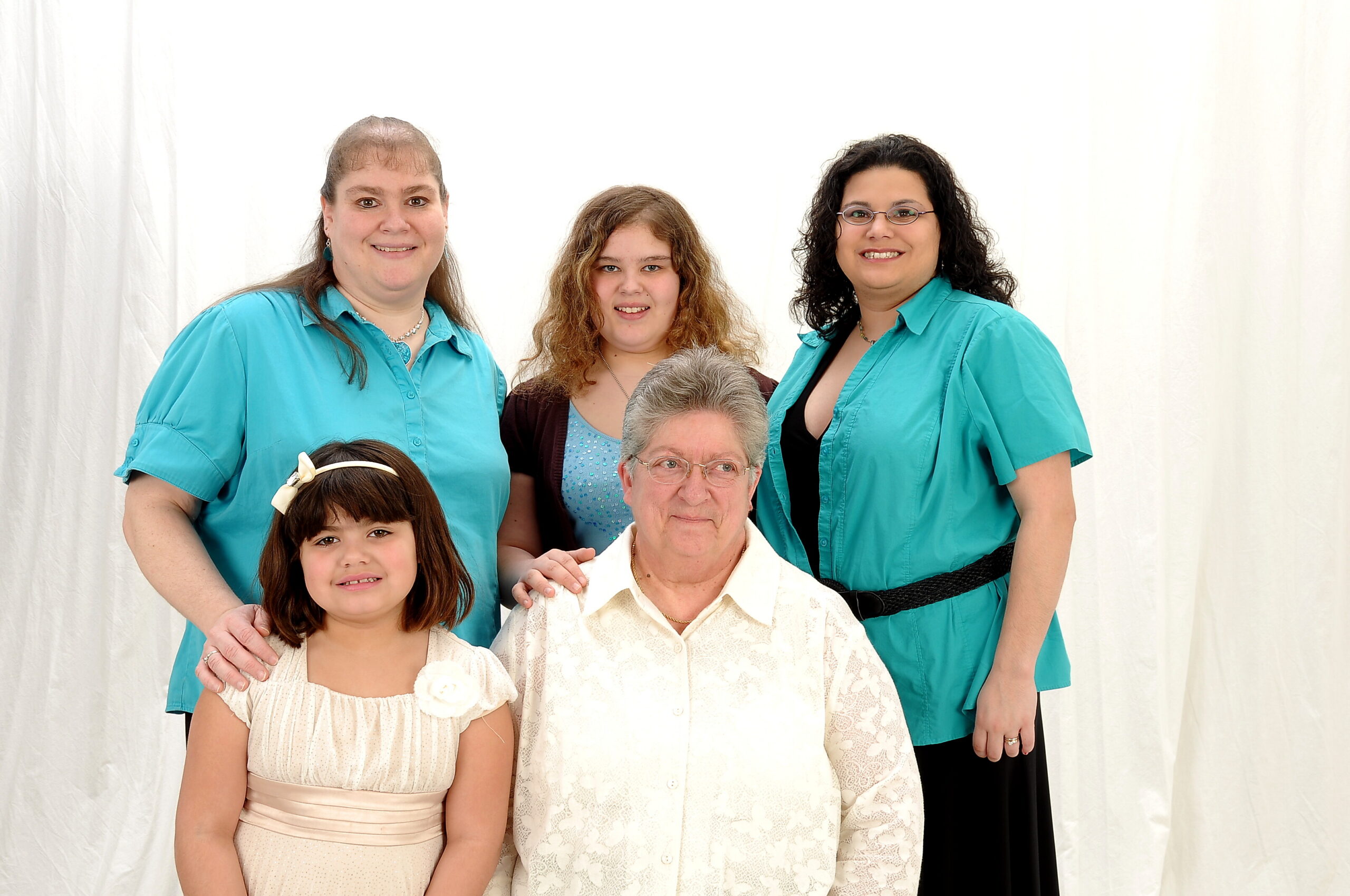 Group portrait of five people standing together: two women in teal shirts on the sides, a young girl in front left, a girl with curly hair center, and an older man seated in front.