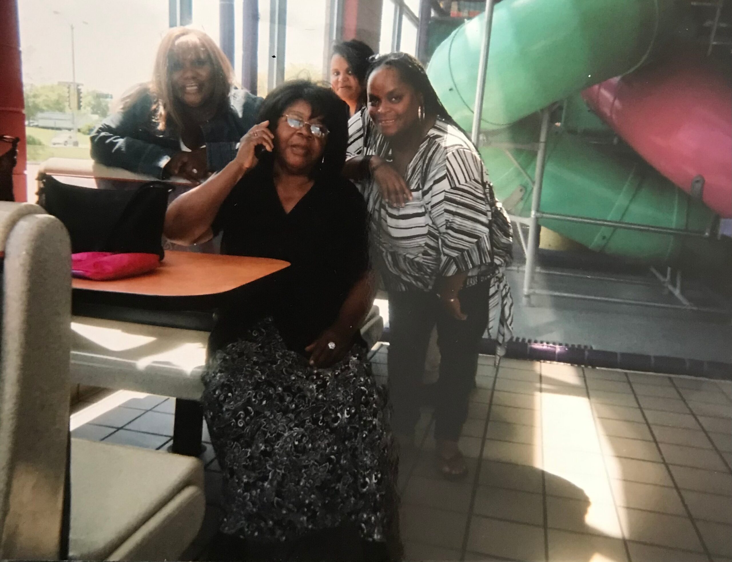 Group of four women posing together indoors; seated woman in black, others stand around her, with a green play structure visible in the background.