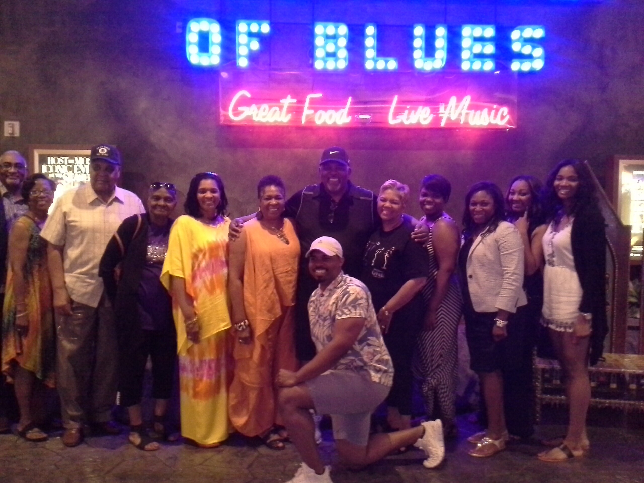 Group of about fifteen people posing for a photo in a venue with neon signs reading 'Great Food - Live Music' above them in blue and pink lights.