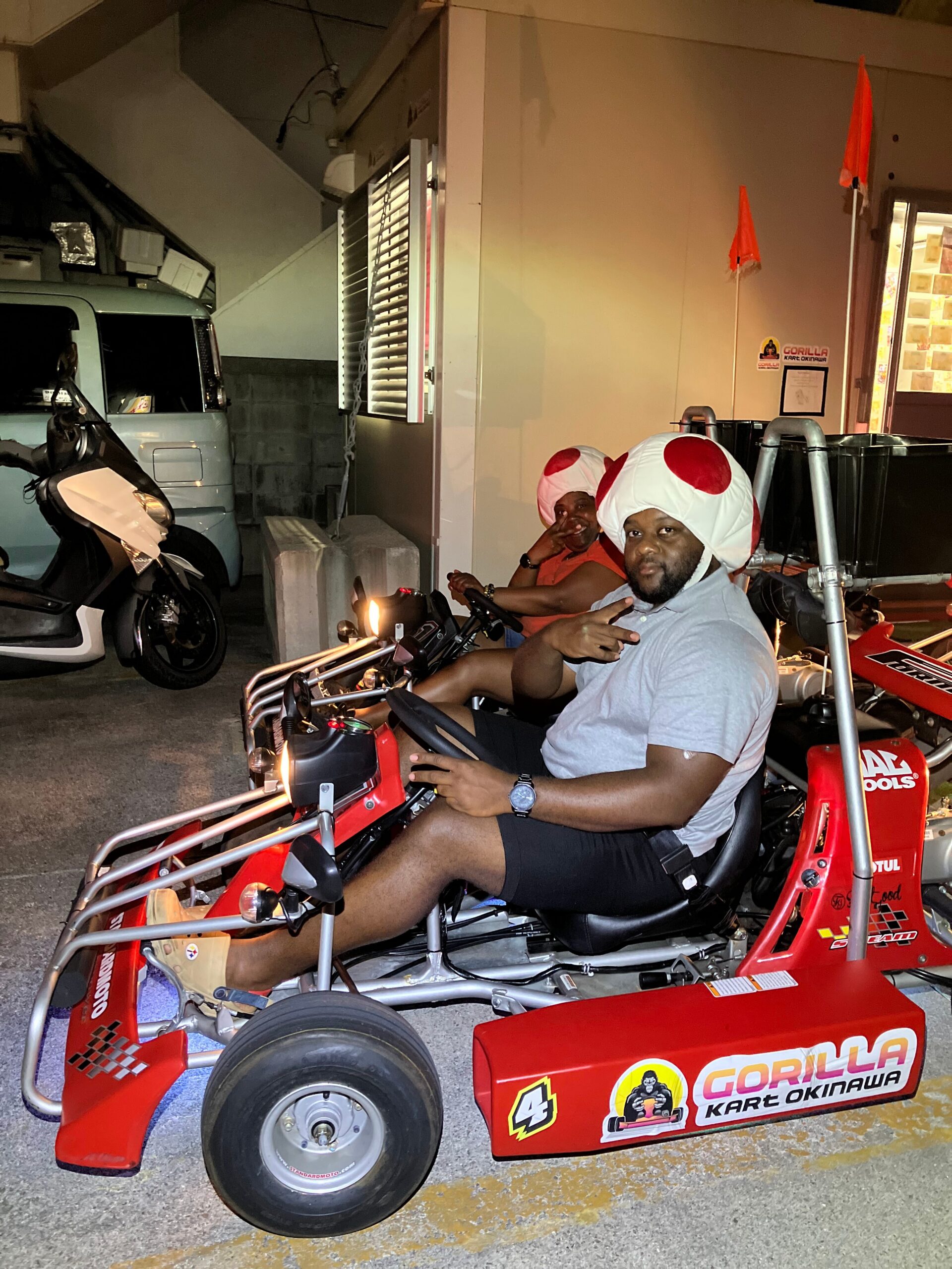 Two men wearing oversized white helmets with red spots sit on red go-karts in a garage, posing for the camera.
