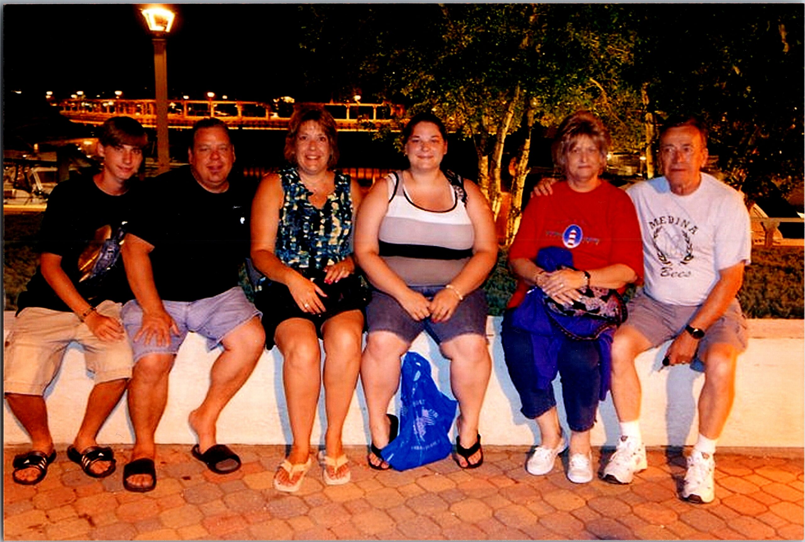Seven adults sit closely on a low wall at night by a waterfront, posing for a group photo with lit trees and a lamp post in the background.