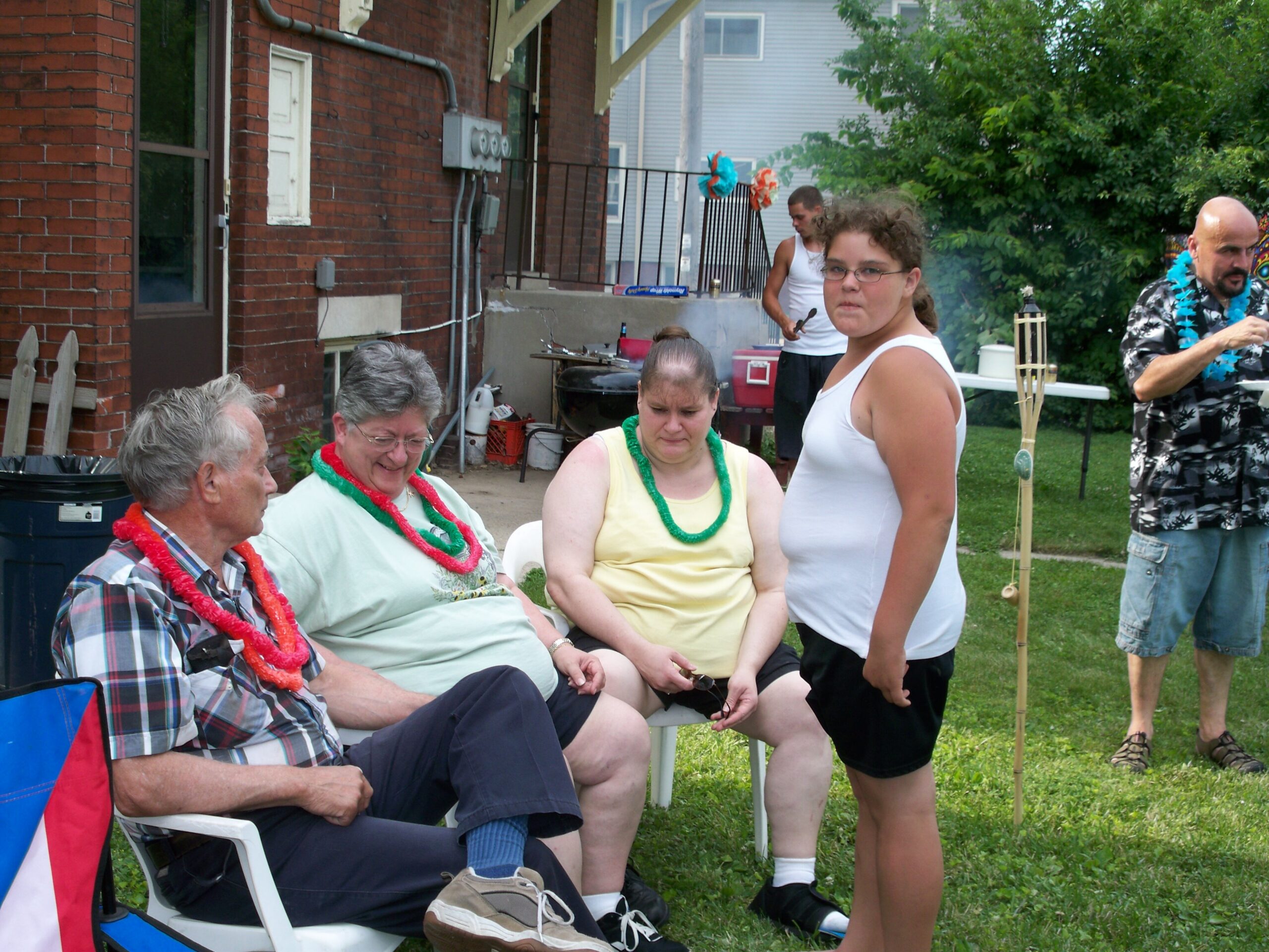 Group of adults at a backyard party; several wear colorful leis, seated and standing near a red-brick building.