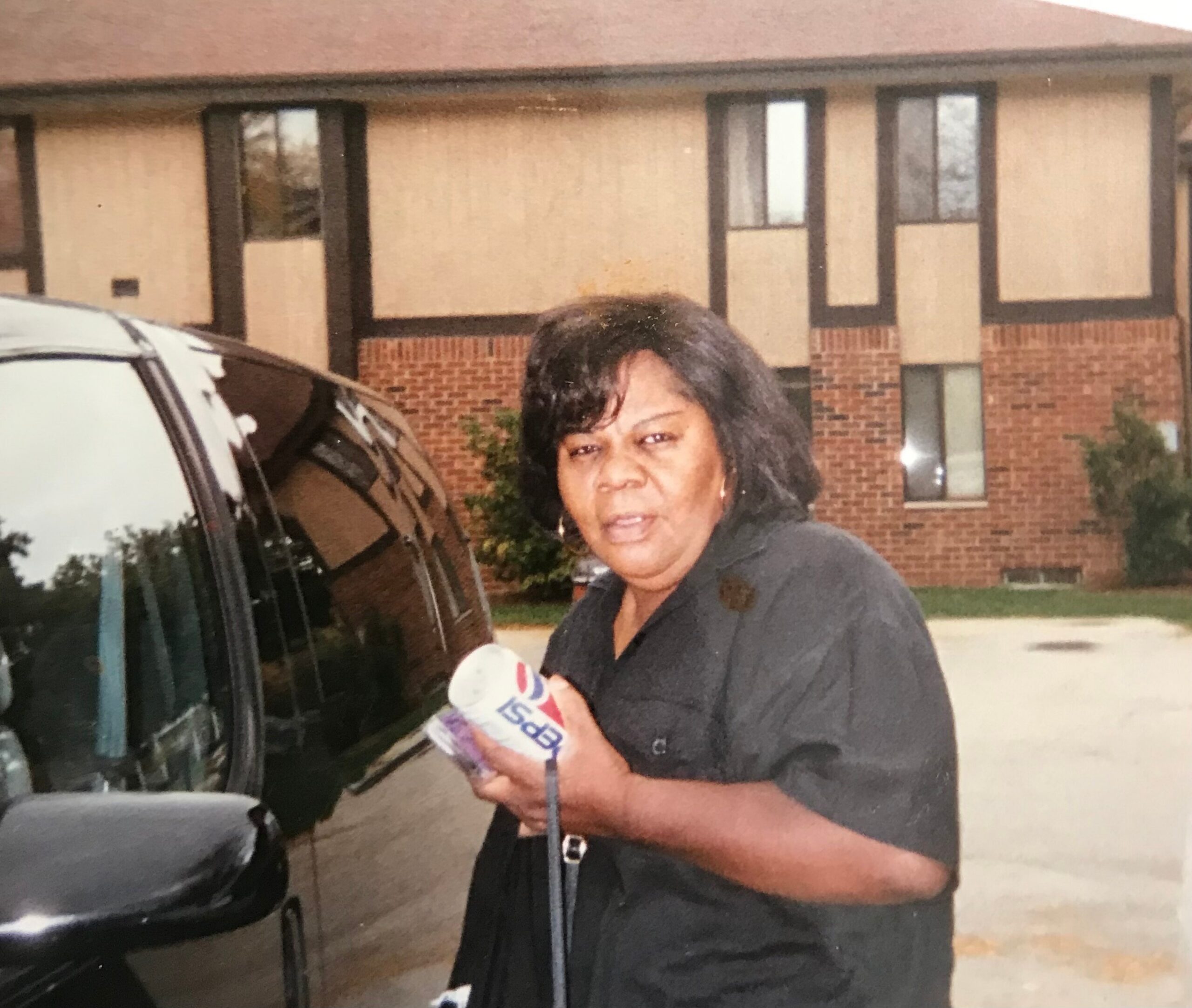 Woman standing beside a dark van outside a brick apartment building, holding a bag of groceries.