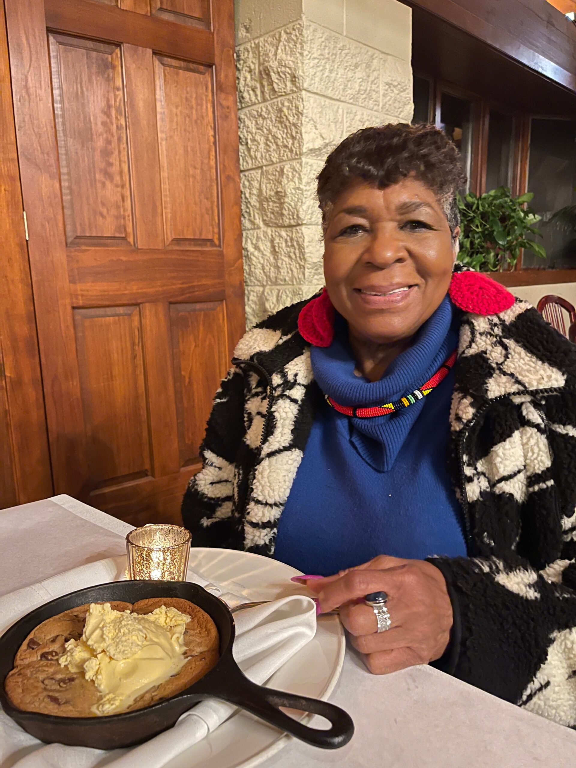 Smiling woman in a blue sweater and patterned coat sits at a restaurant table with a cast-iron skillet dessert topped with ice cream in front of her.