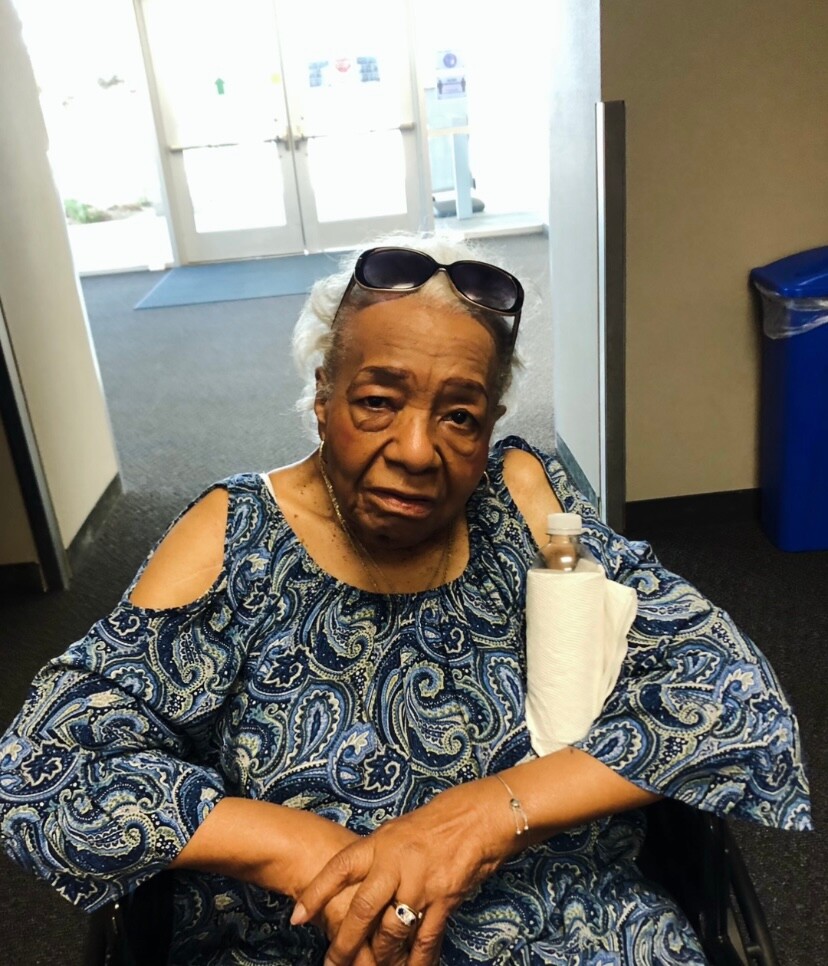 Elderly woman in a wheelchair wearing a blue paisley top, sunglasses on her head, sitting in a building lobby.