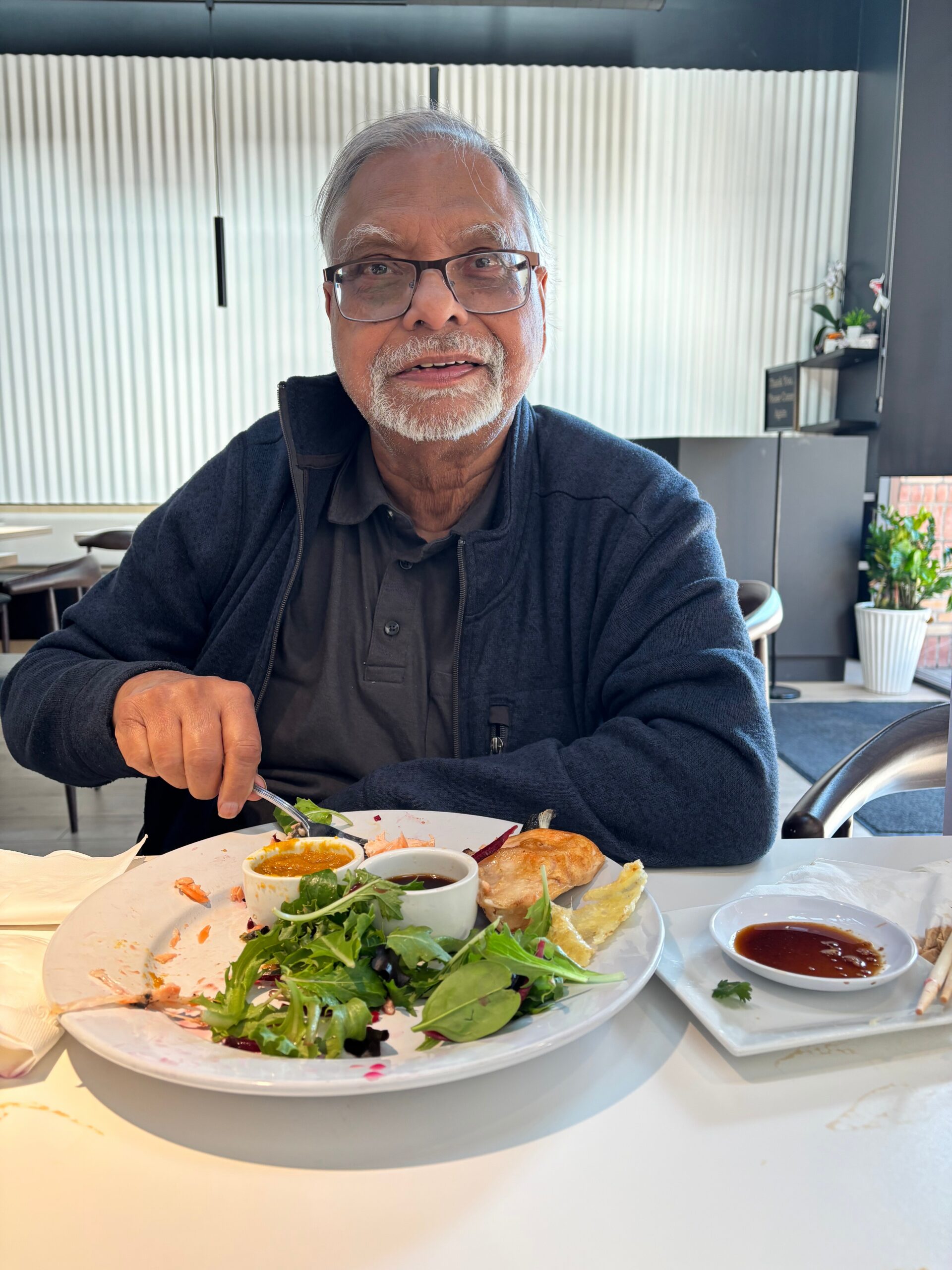 Older man with glasses and gray hair sits at a restaurant table, eating salad with sauces and bread visible on the plate in front of him as he looks at the camera/screen.