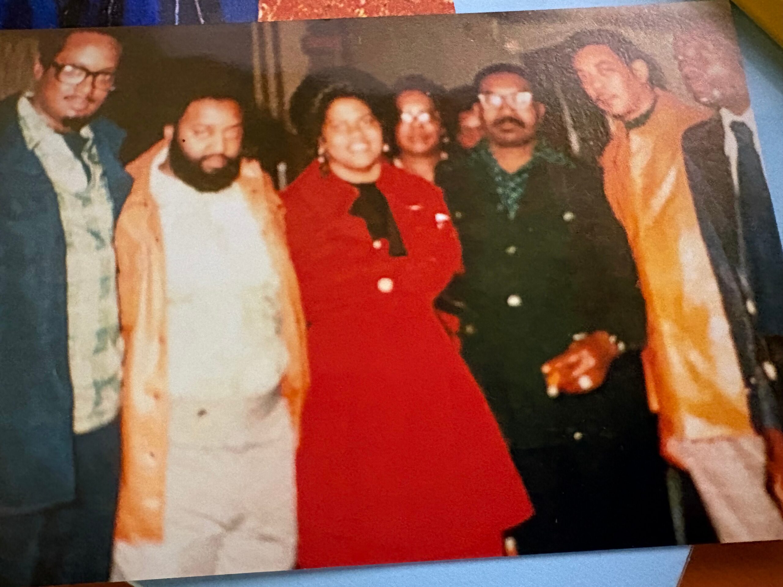 Group of people posing closely together at an indoor party, smiling for the camera (center figure in a red outfit).