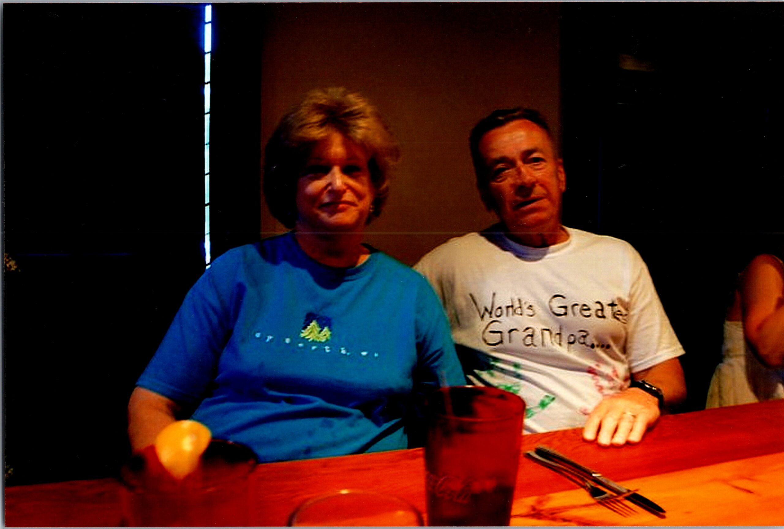 Two adults sit at a wooden table with drinks, smiling for the camera (woman in blue shirt left, man in white shirt right).