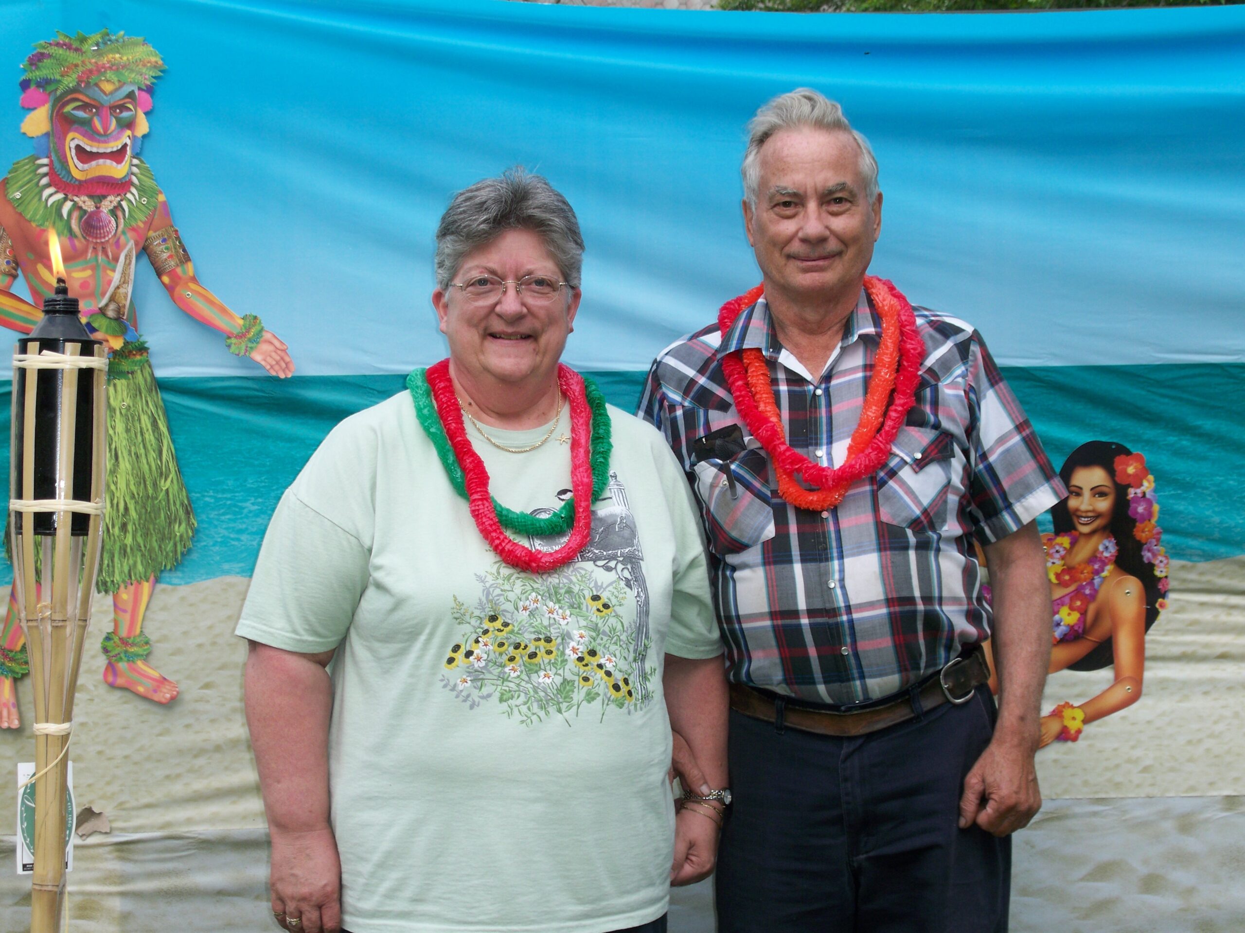 Two smiling adults wearing colorful leis pose in front of a tropical backdrop at a beach-themed event.