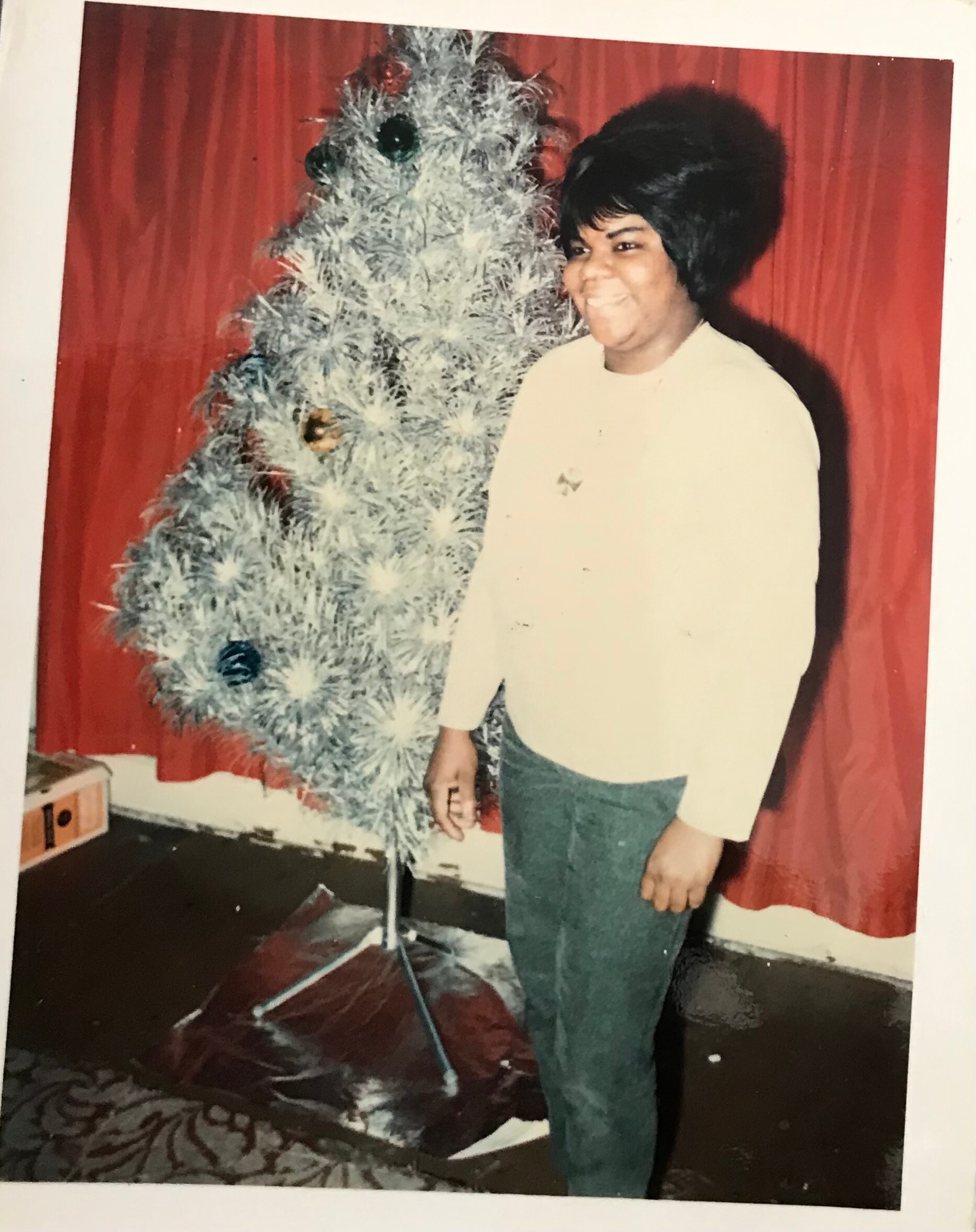 Person standing beside a silver Christmas tree indoors, wearing a light shirt and jeans, with red curtains in the background.