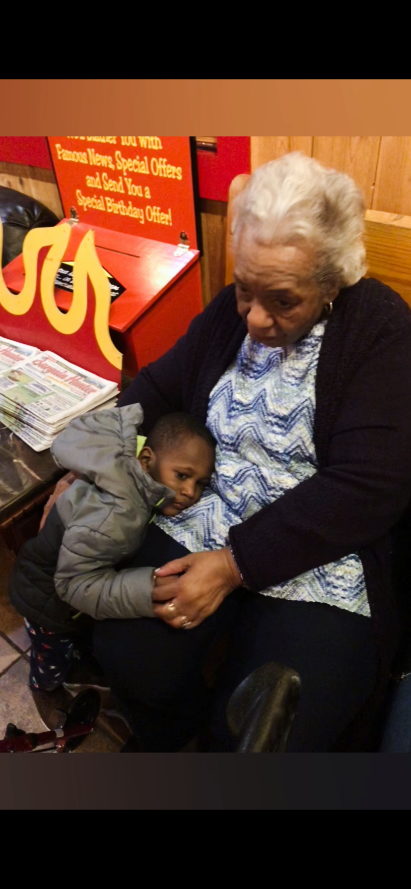 Grandmother sits with a young child resting his head on her lap in a shop, with newspapers on the counter nearby.