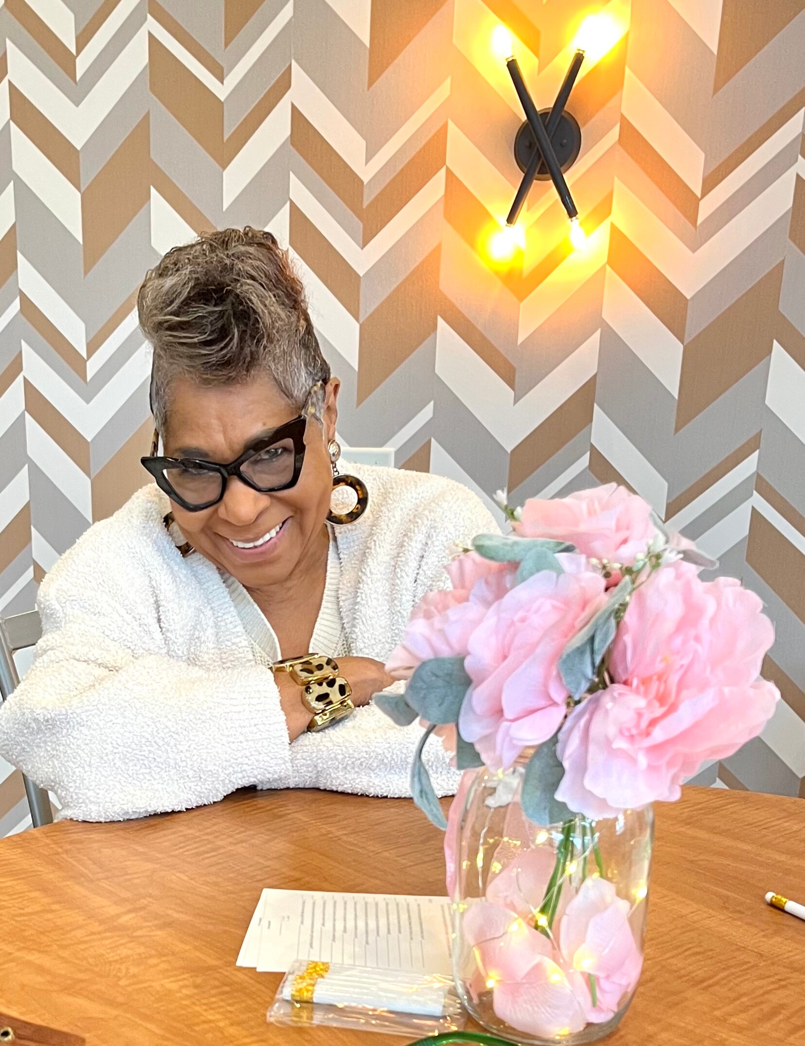 Smiling woman with short gray hair and black glasses sits at a wooden table with a pink bouquet in a vase behind her; geometric beige wallpaper in the background.