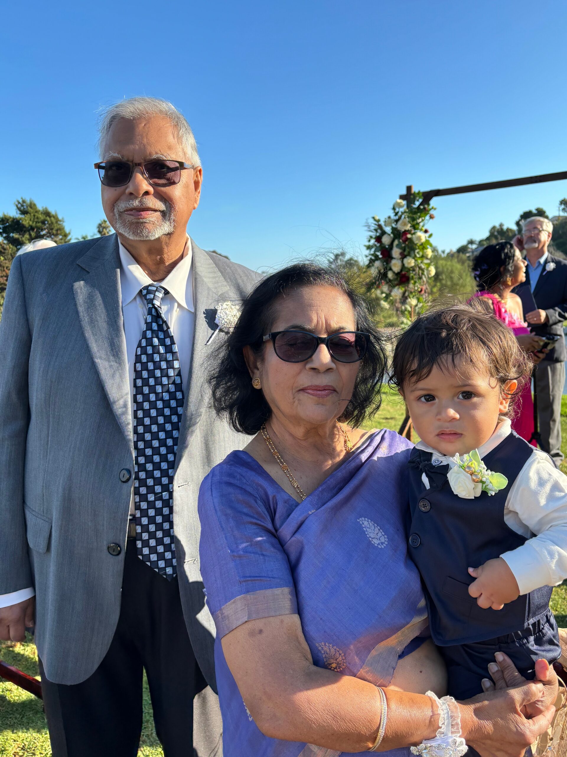Older couple with a toddler at an outdoor wedding; man in a gray suit and patterned tie, woman in purple holding the child, wedding arch in background.