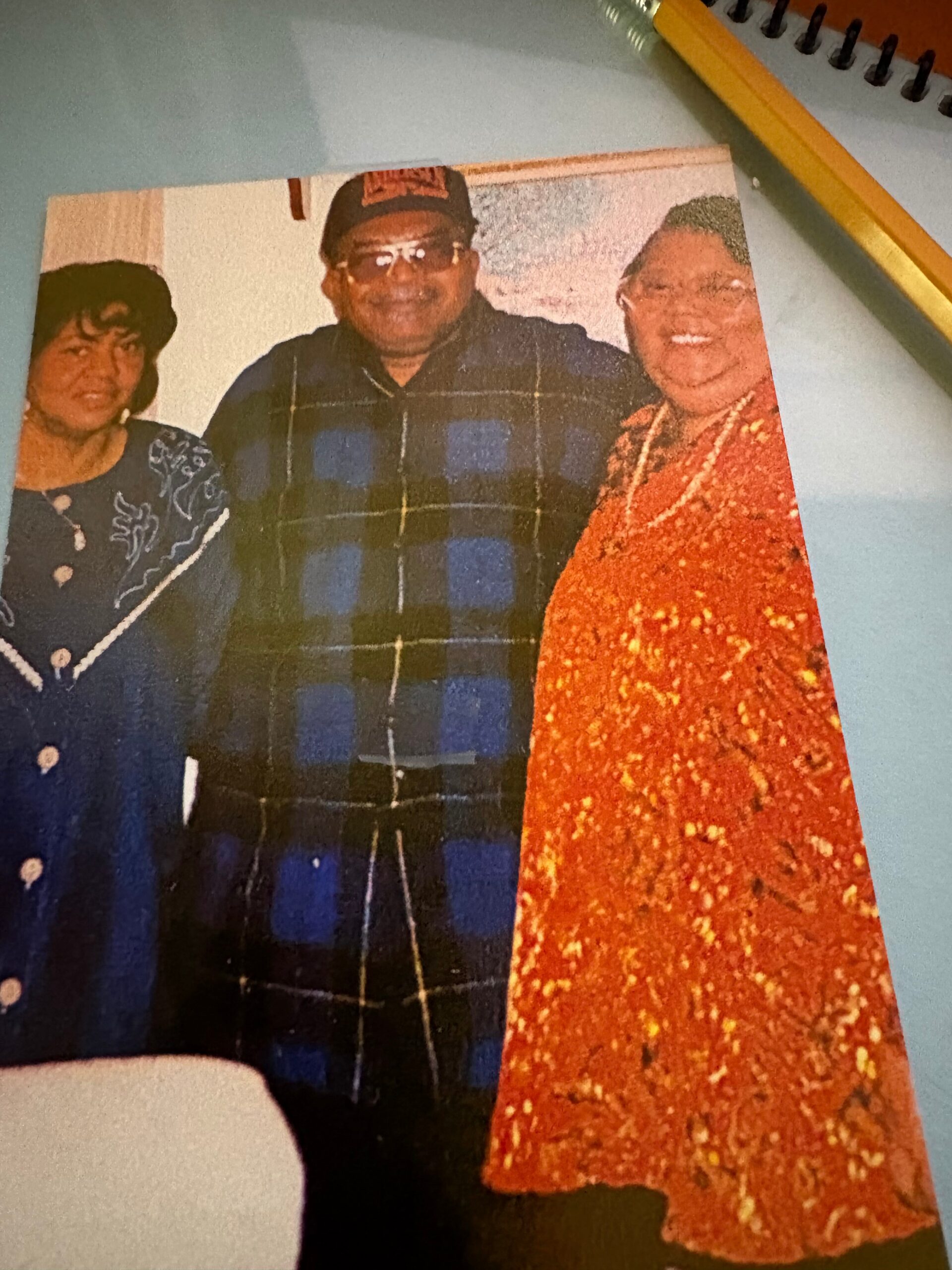 Three adults posing for a family photo: woman in blue-embroidered outfit, man with cap and glasses in the center, and a woman in a bright orange sparkly dress on the right.