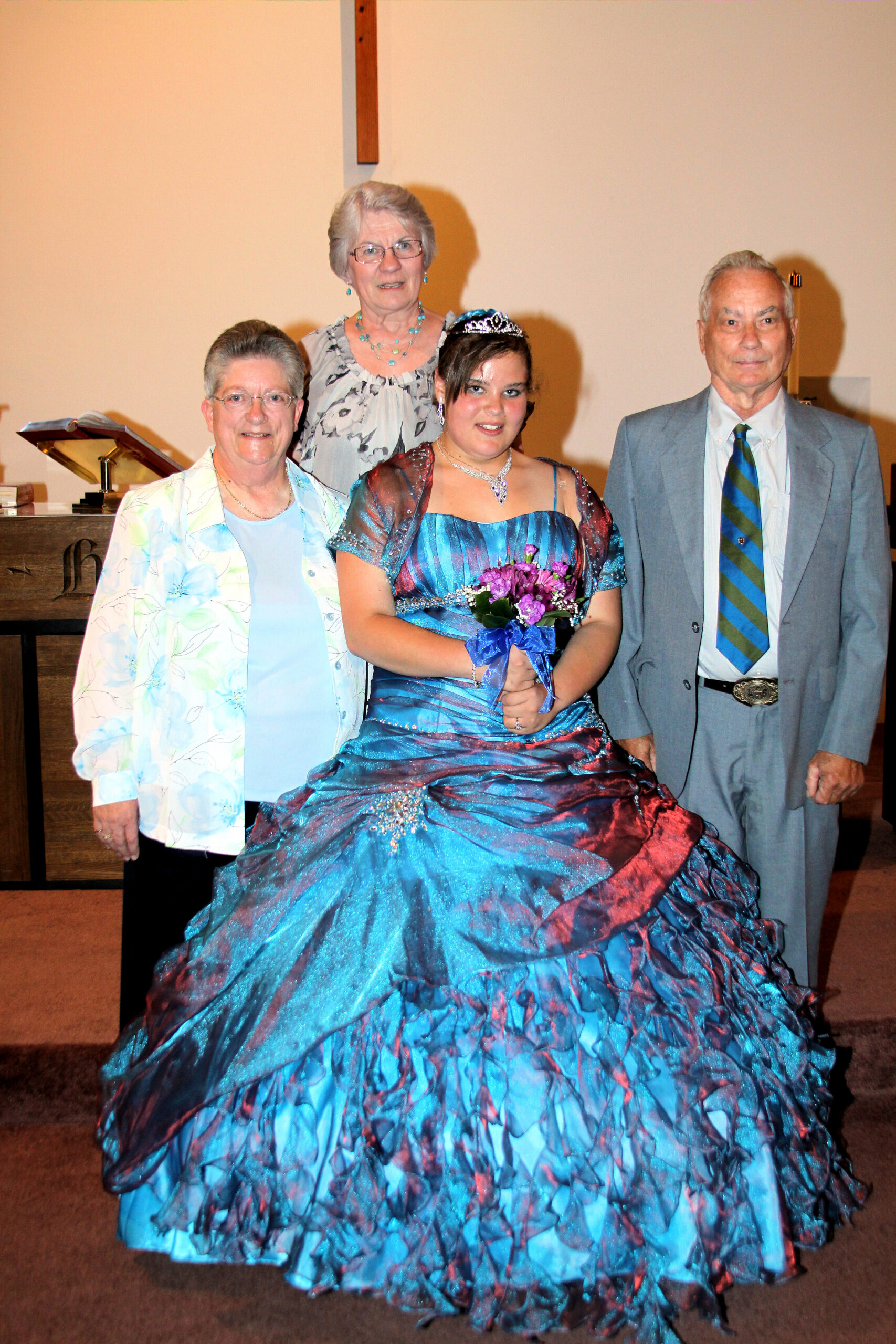 Young woman in a blue ballgown with a tiara and bouquet posing with two older women and a man at a formal event. In a church-like setting with a cross in the background, onlookers nearby.