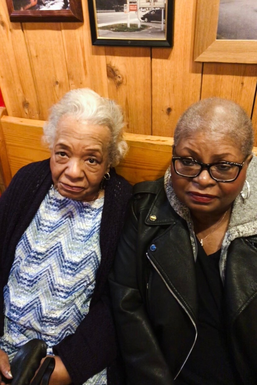 Two elderly African American women sitting side by side in a wood-paneled room, looking at the camera.
