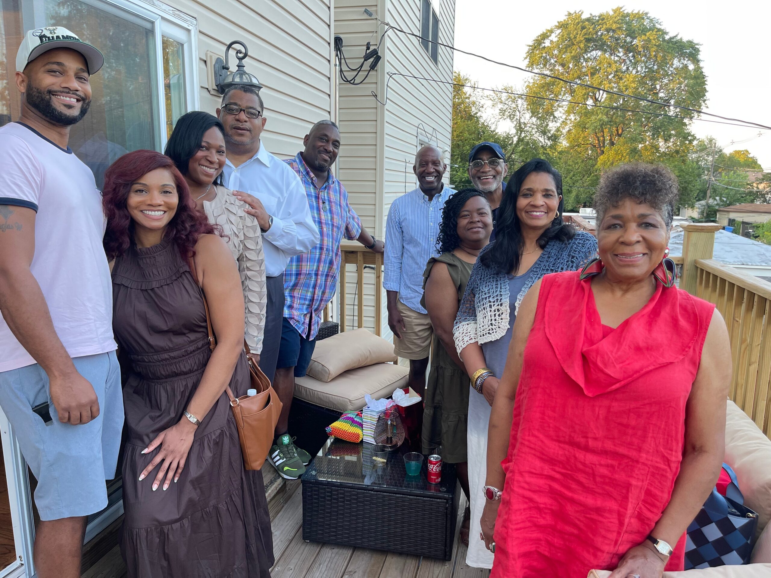 Group of adults standing on a wooden deck beside a house, smiling for a group photo.