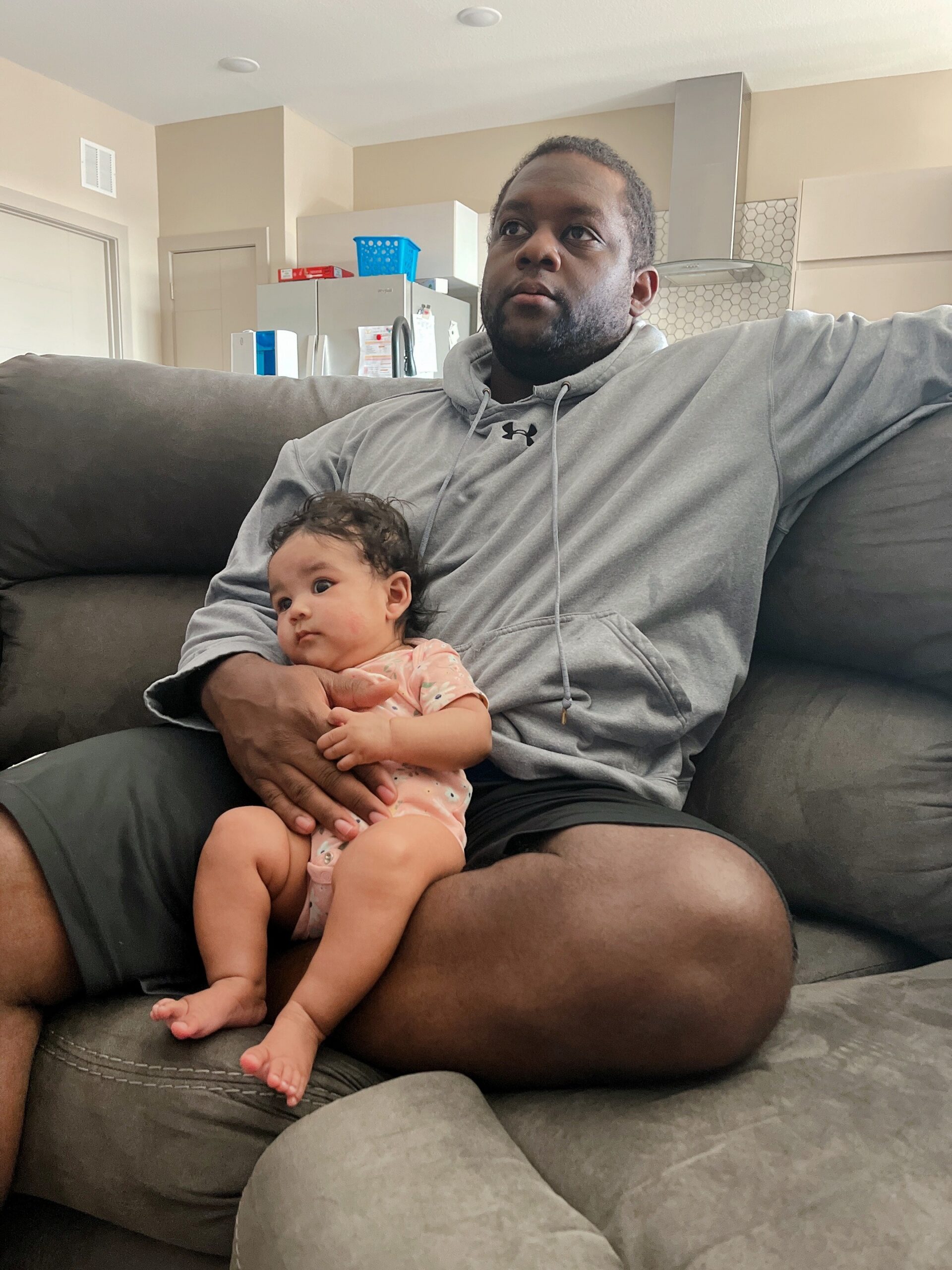 Man in a gray hoodie sitting on a couch, cradling a baby in a pink baby onesie.