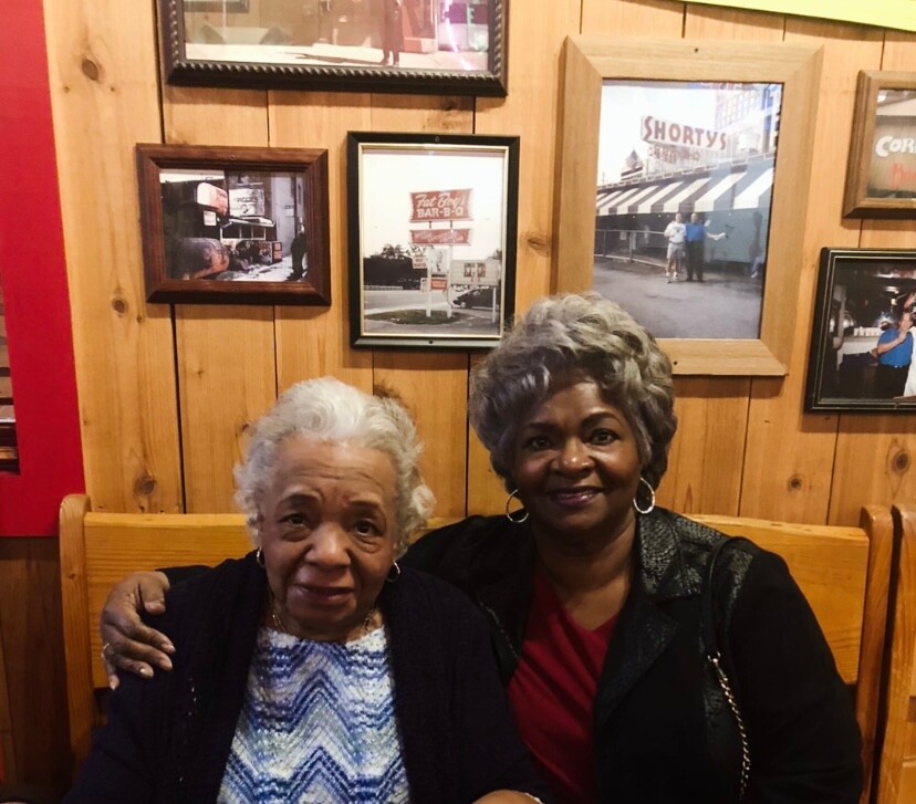 Two smiling elderly African American women sit close together, arms around each other, on a wooden bench with framed photos on a wood-paneled wall behind them.