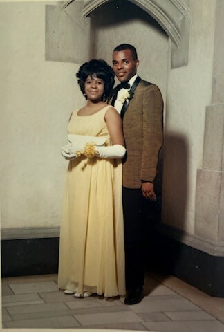 Young couple posing in a formal setting; woman in a pale yellow gown with white gloves and a corsage, man in a brown blazer with a boutonniere.