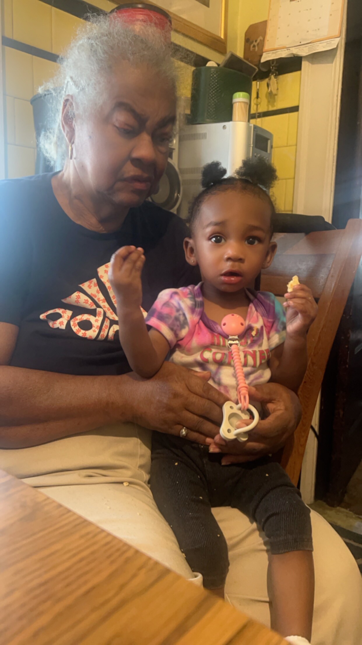 Grandmother with gray hair sits with a toddler on her lap at a dining table, the child holding a small snack and a pink pacifier clip.