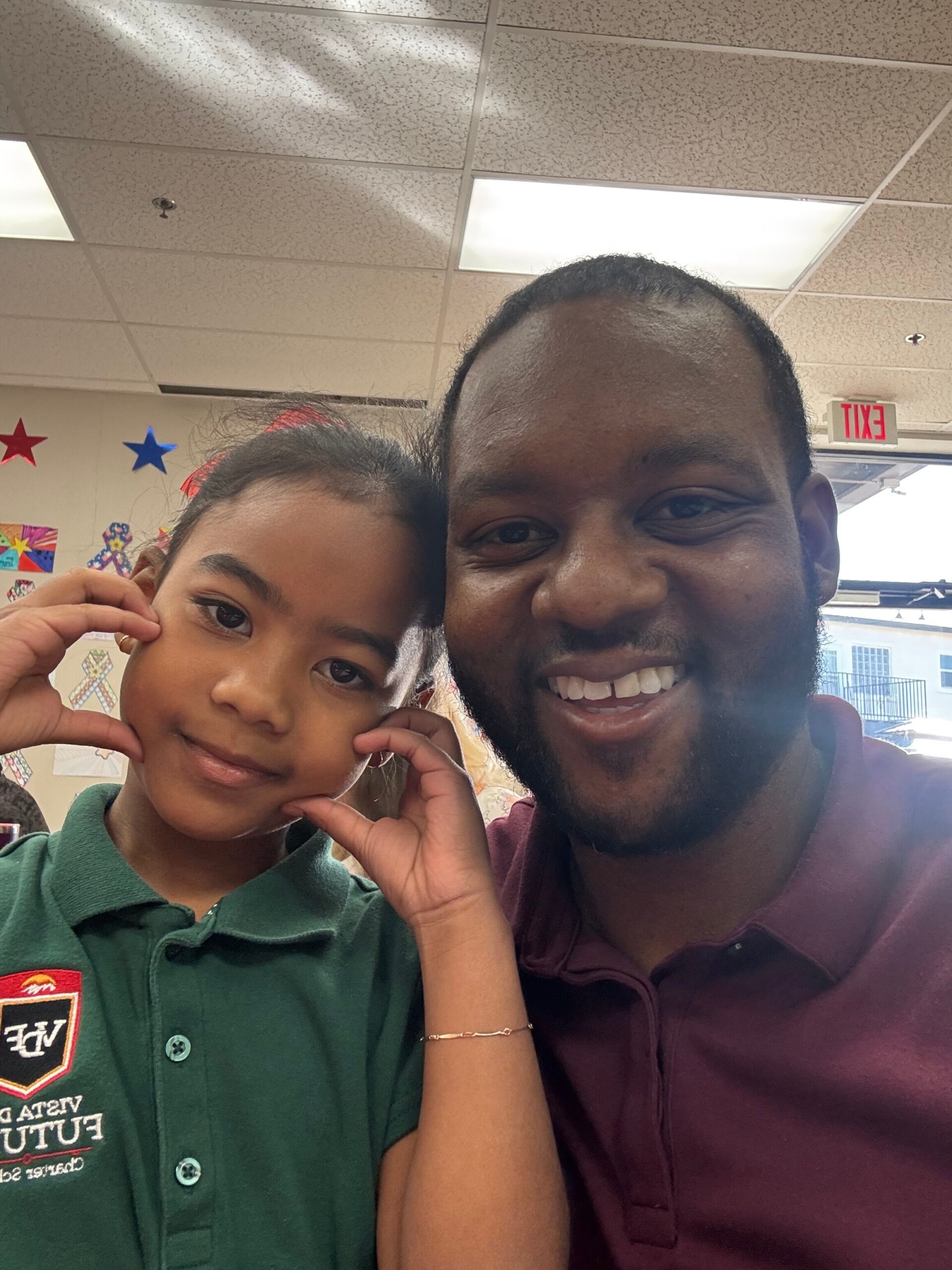 Smiling man and girl posing for a selfie in a classroom with colorful decorations in the background (exit sign visible).